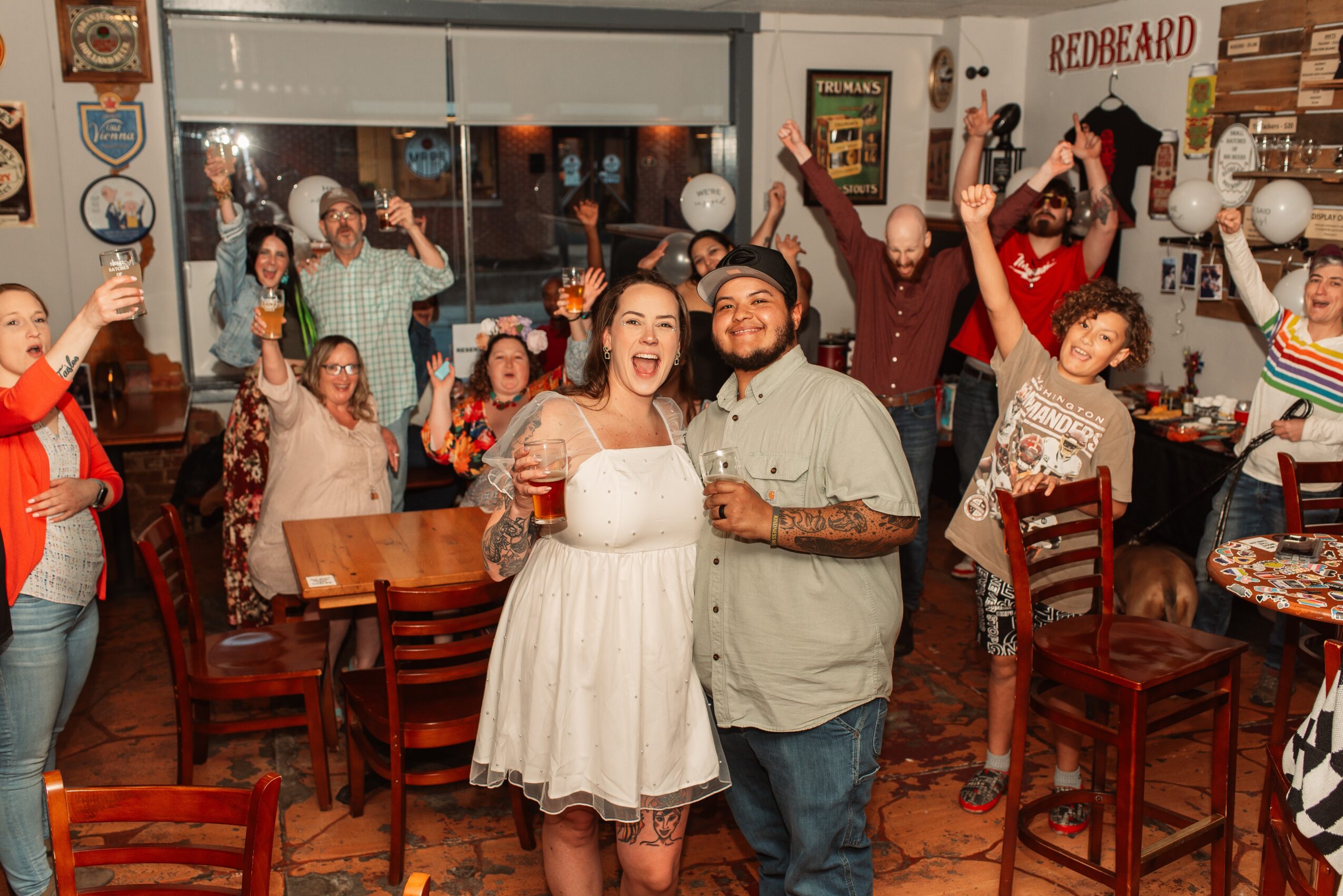People celebrating indoors, some holding drinks, smiling, and raising hands, with a couple in the center, a woman in a white dress and a man with a beard, surrounded by friends.