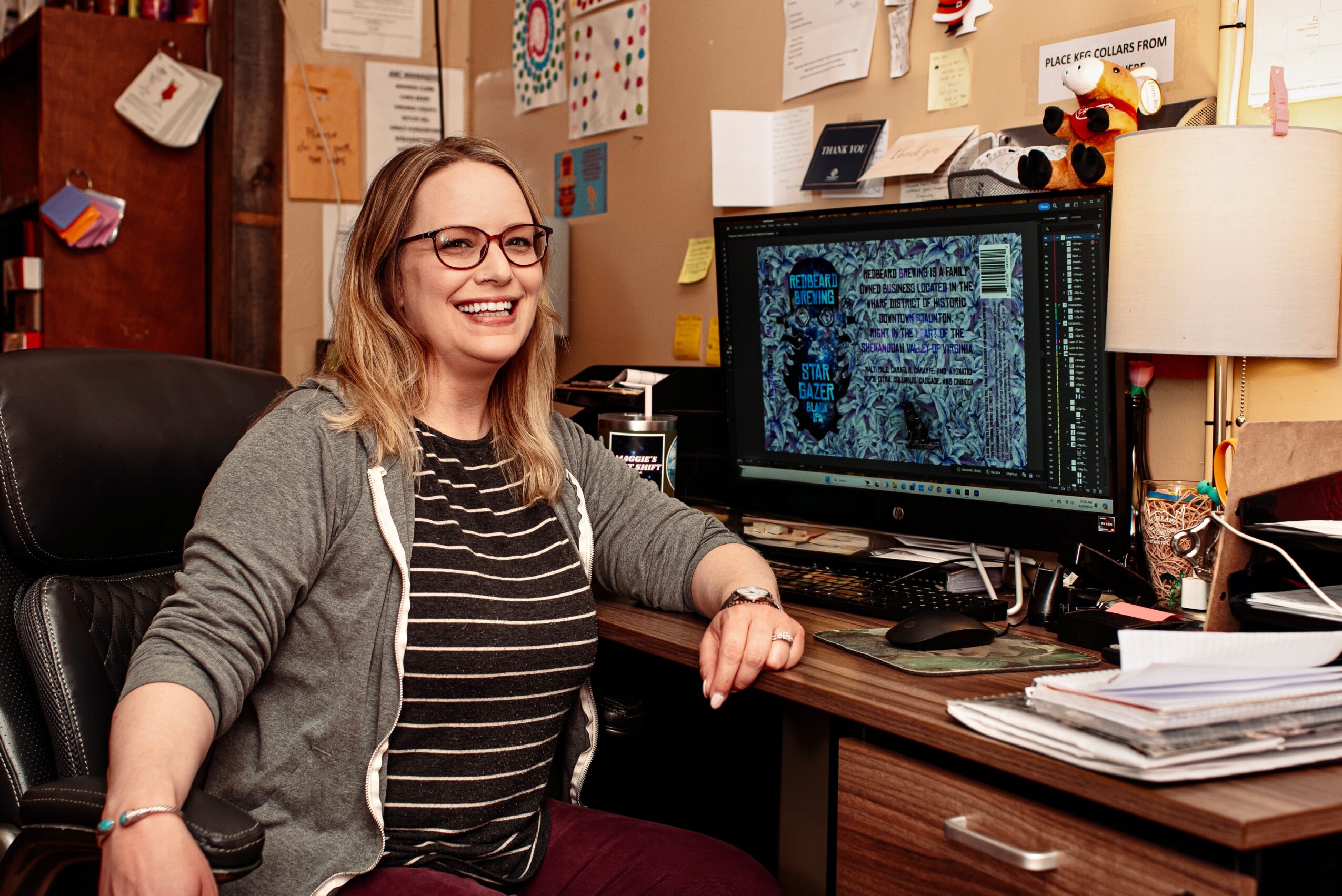 Woman with glasses smiling at computer in cluttered office, papers and bulletin board behind her.