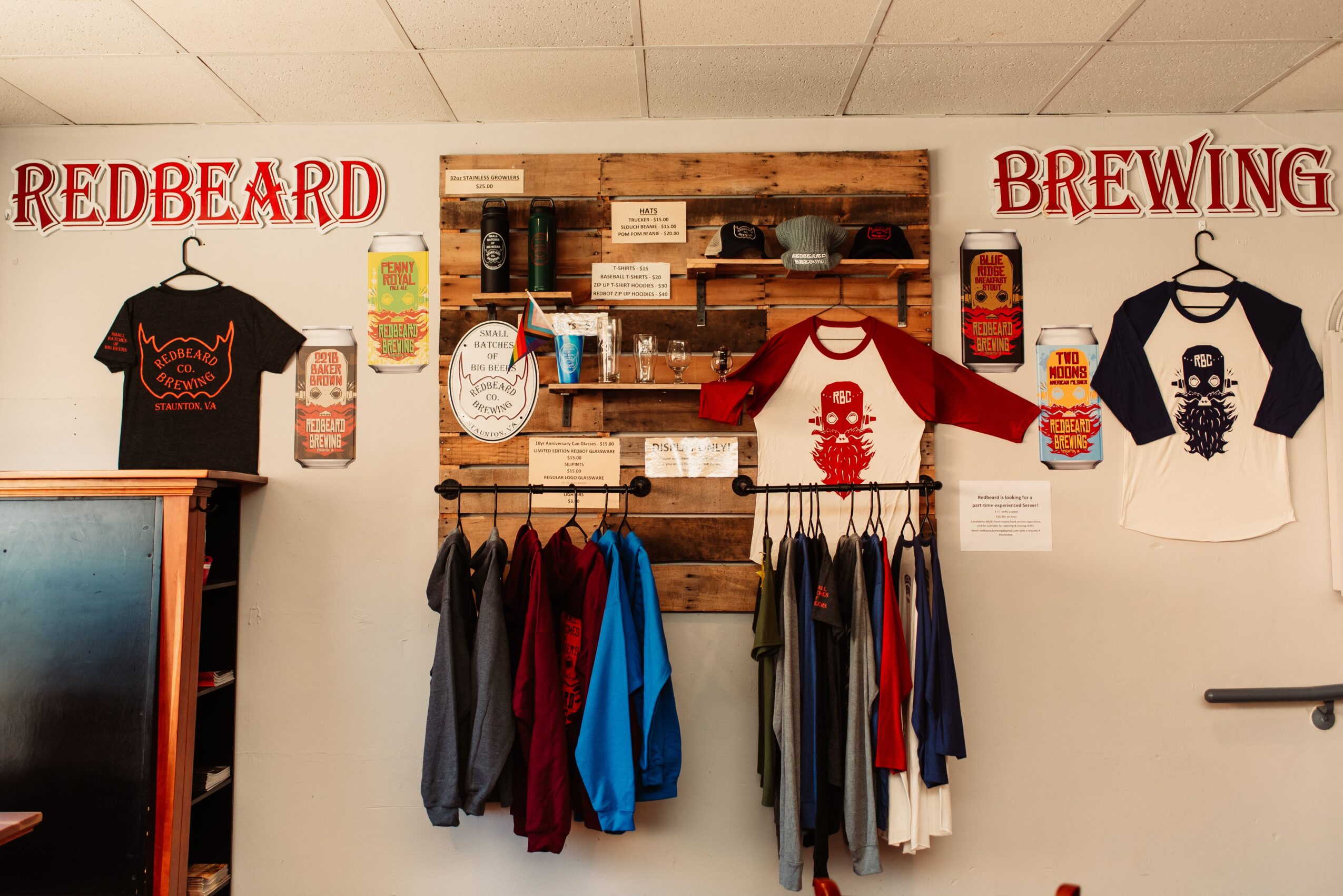 Display of T-shirts, clothing, and brewing-themed decorations on a wall with wooden shelves, with signs reading 'REDBEARD' and 'BREWING'.