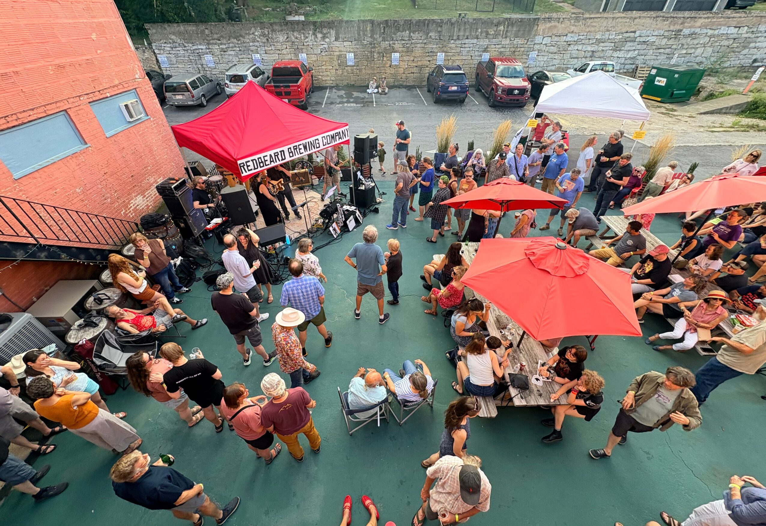 People gather outdoors under umbrellas and tents, socializing and eating at a community event on a green surface.