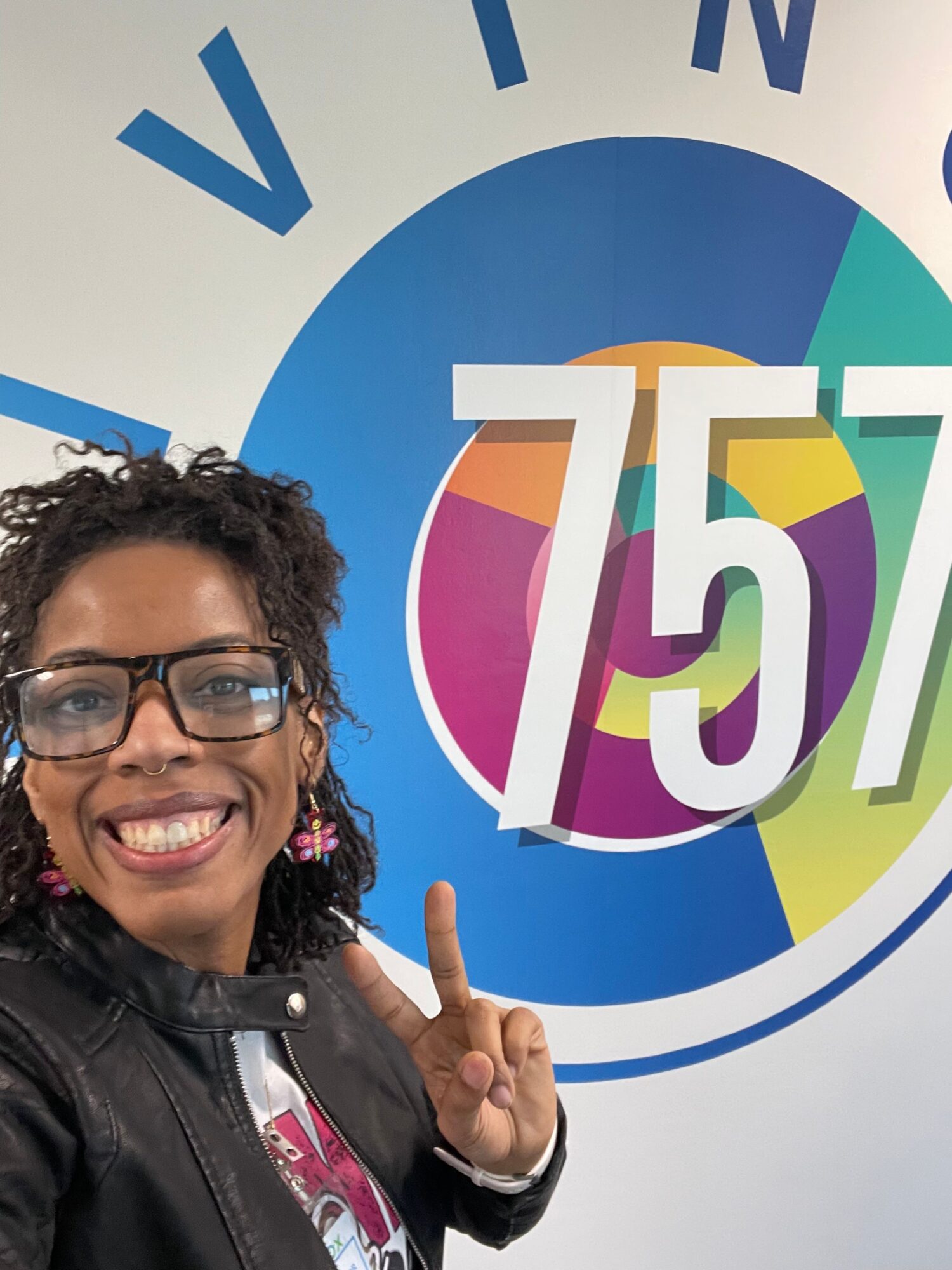 Smiling woman with glasses and curly hair pointing at a colorful circular logo on a wall.