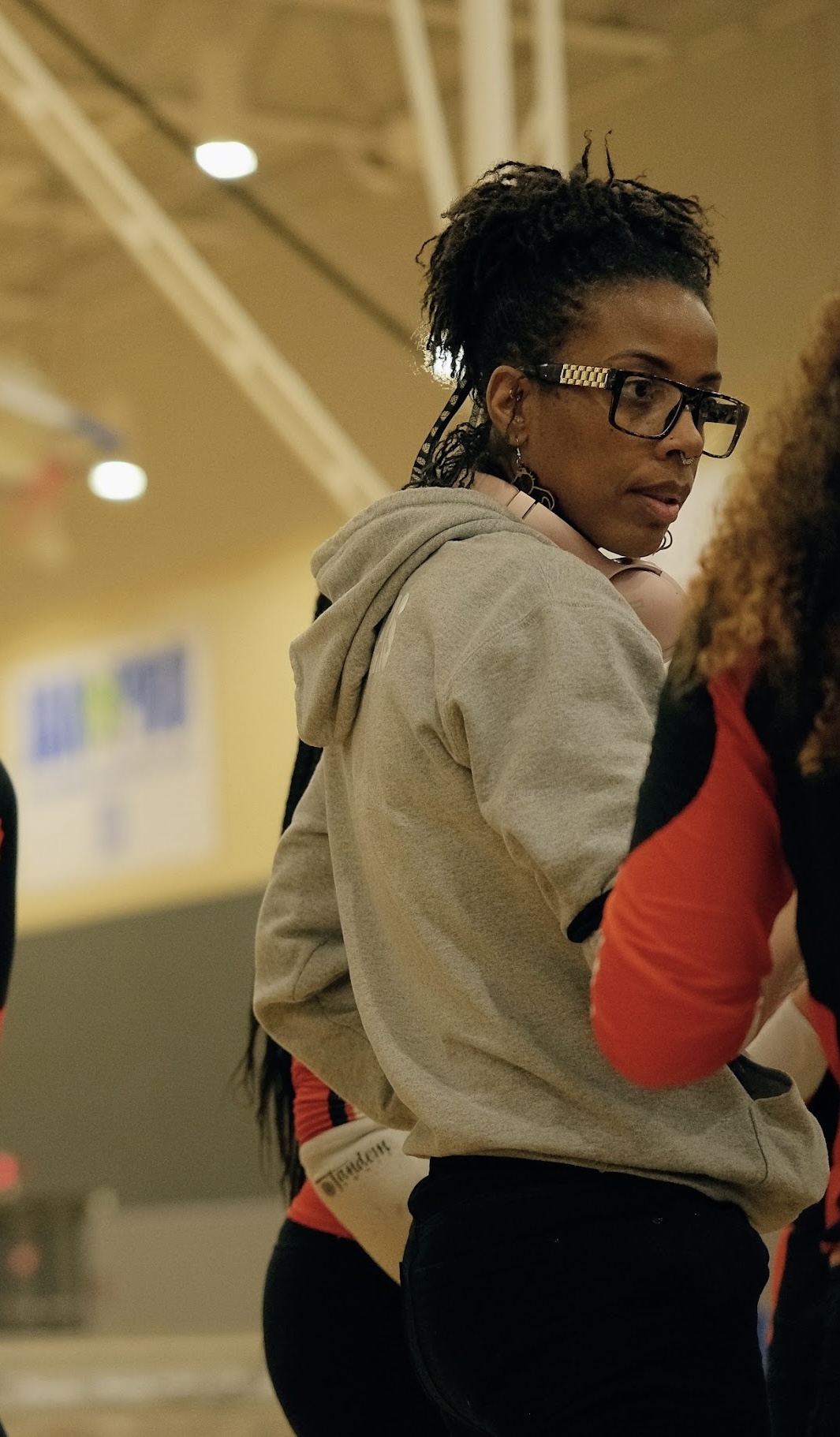 Young woman with glasses and braided hair wearing a gray hoodie, standing indoors with a yellow background.