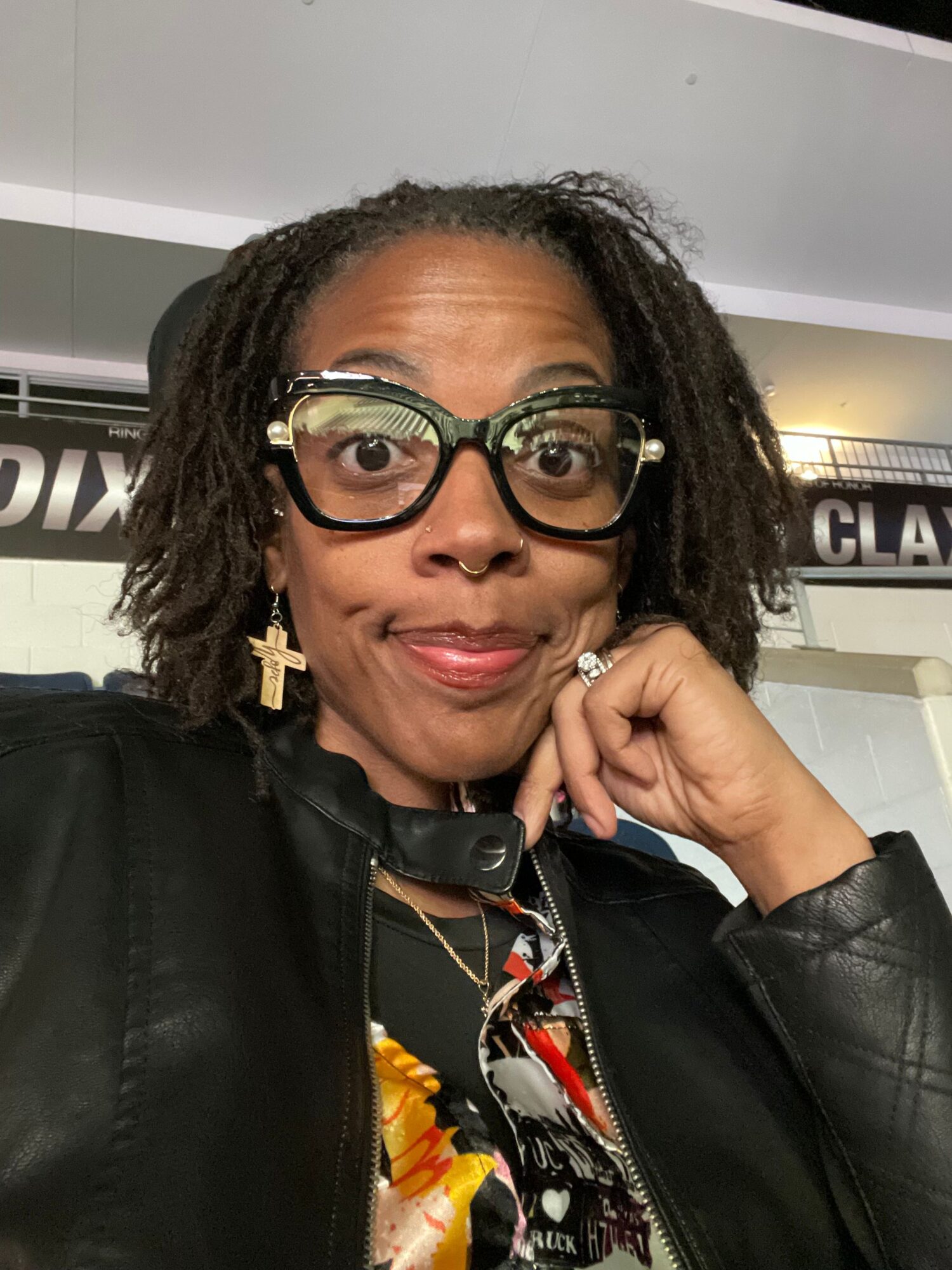 Woman with glasses and earrings, wearing a black leather jacket, resting her face on her hand.
