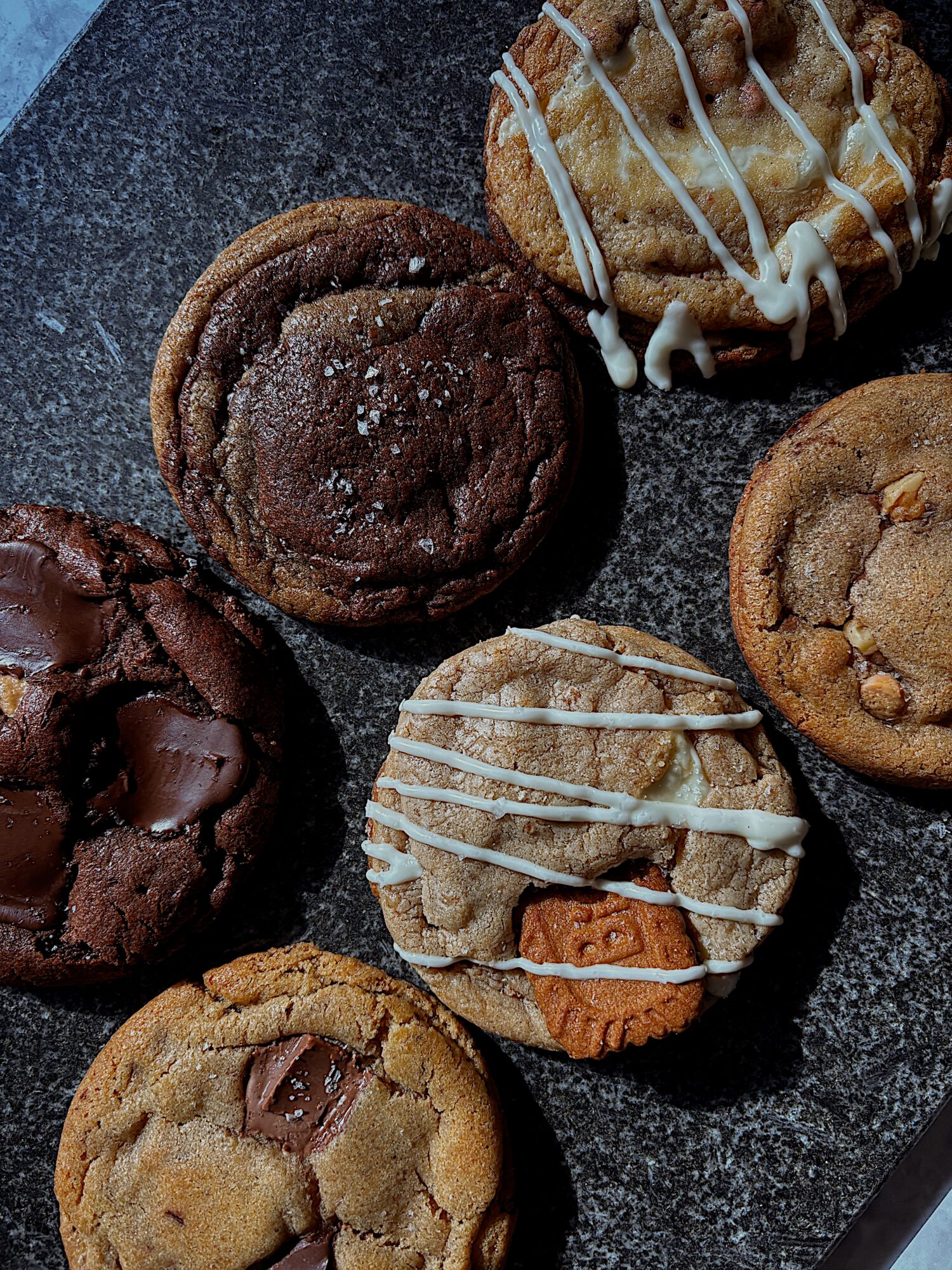 Six cookies on a dark surface, with various flavors and toppings, including chocolate, white icing, and nuts.