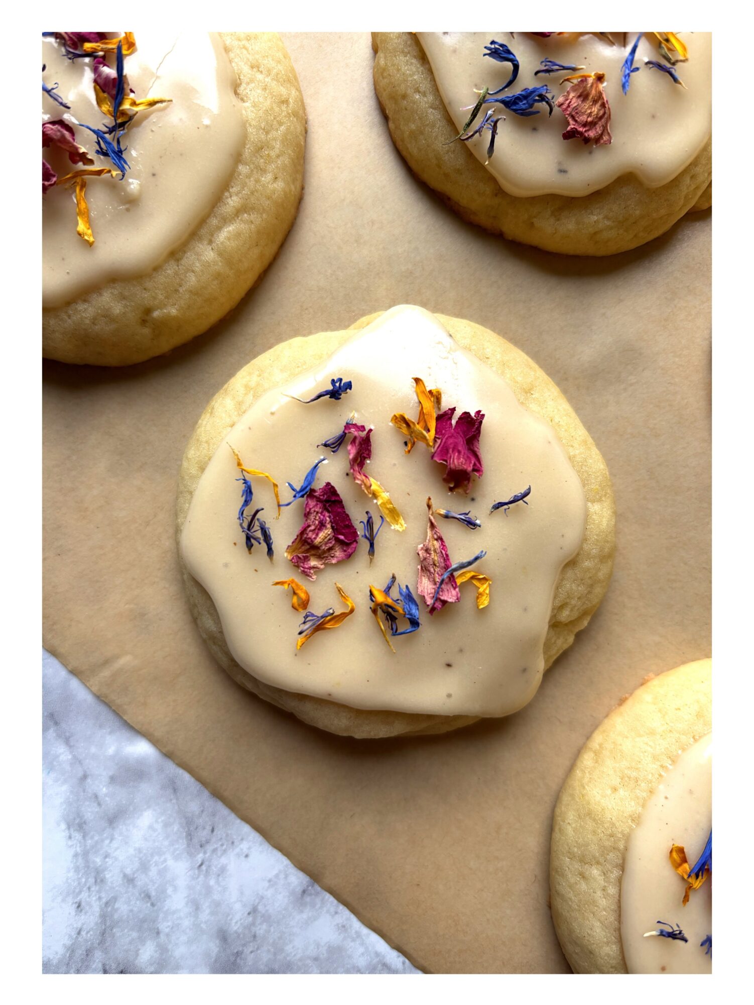 Cookies with white icing and colorful edible flower petals on top, on parchment paper.
