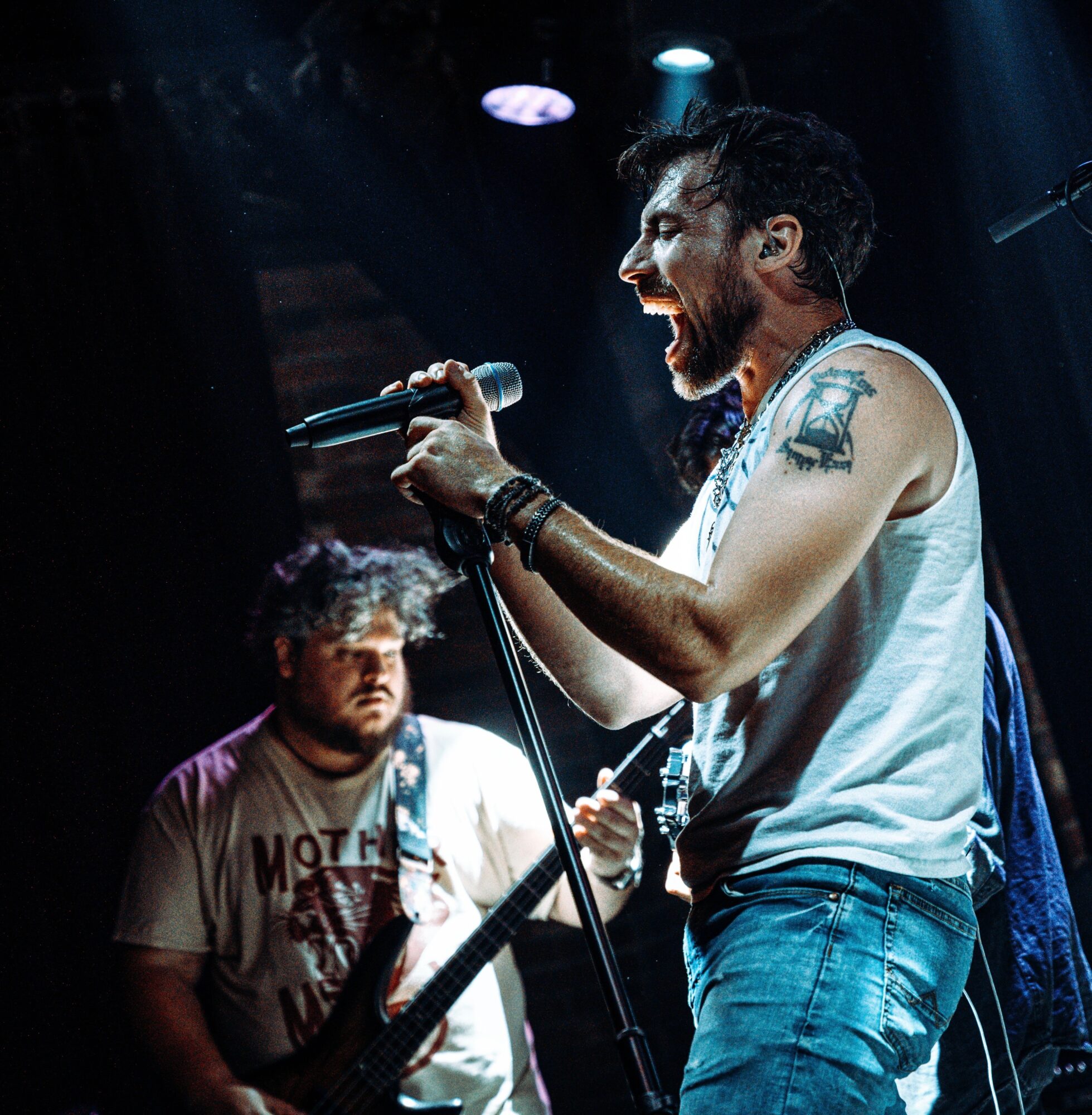 Male singer passionately performs with microphone, wearing sleeveless shirt, tattoos, and jeans, with a guitarist in background, stage lighting visible.