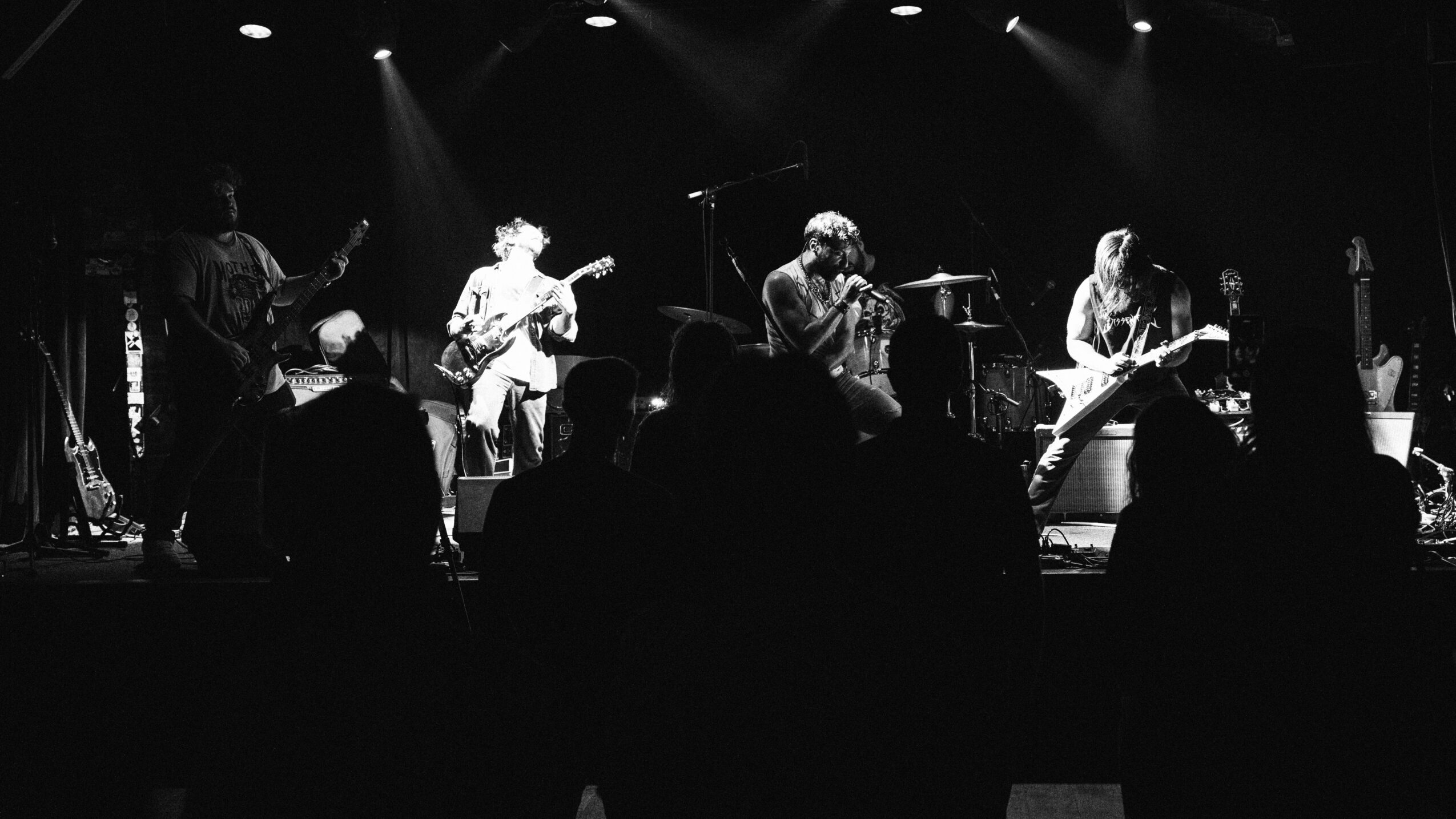 Band performing on stage with audience in foreground, stage lights shining down, black and white image.