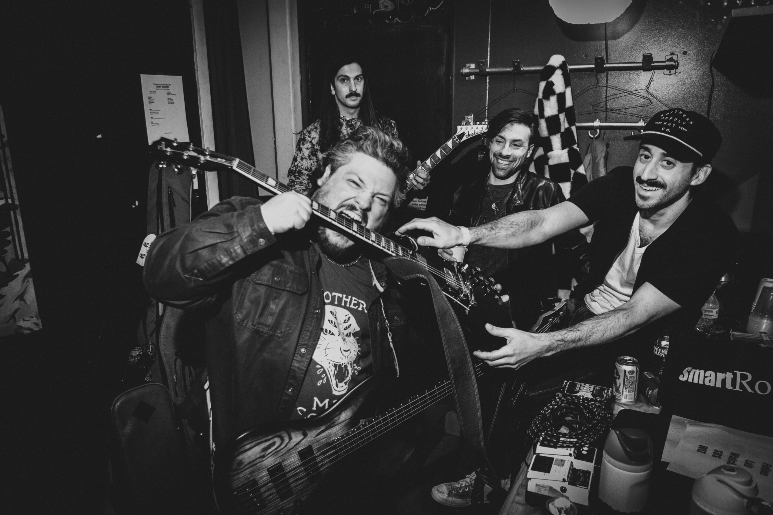 Four men in a room, one playing guitar, others reaching out, with musical equipment and a checkered cloth in the background.