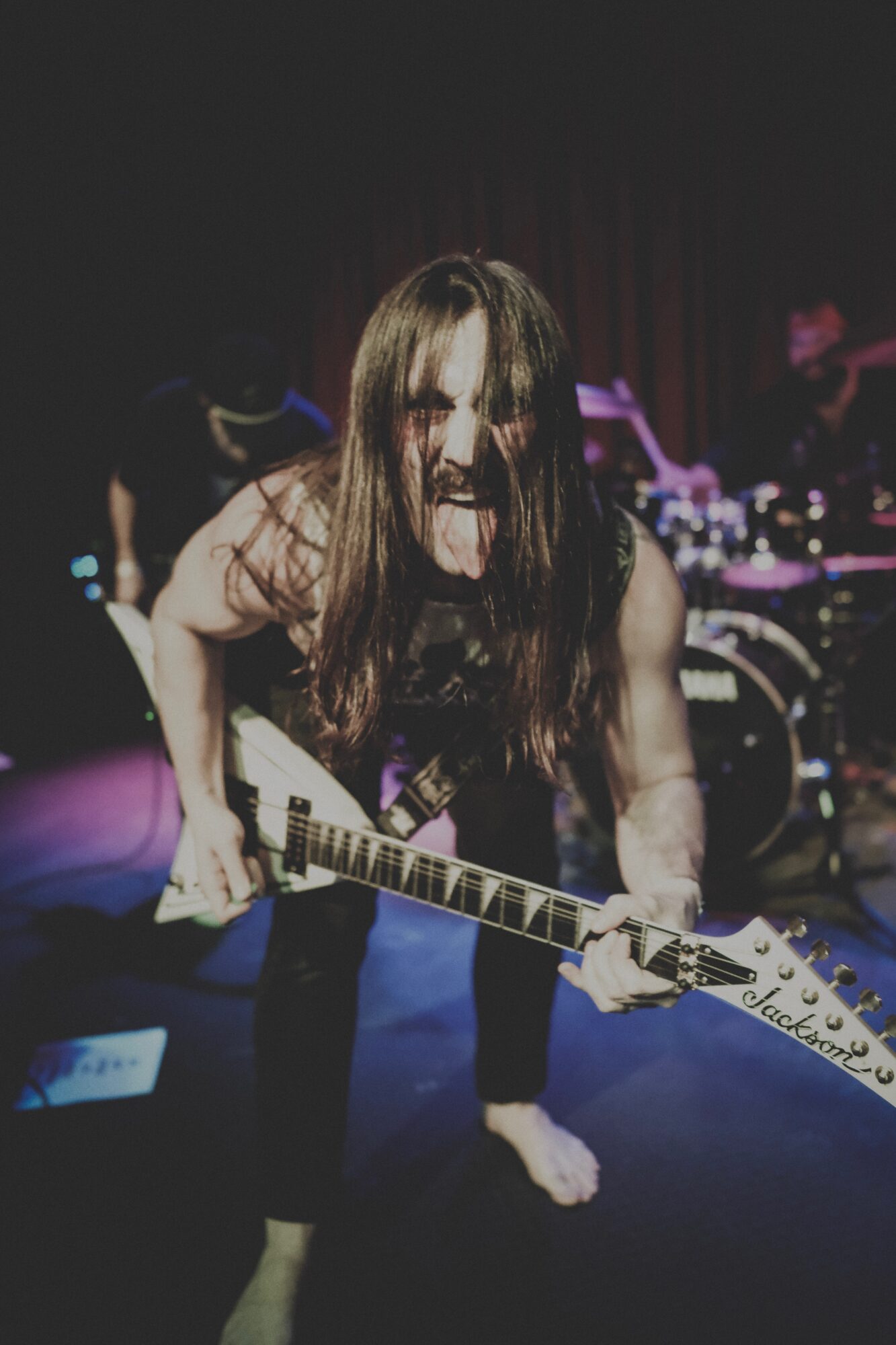 Female guitarist with long hair playing guitar on stage, sticking out tongue, dark background, stage lights, band members in background