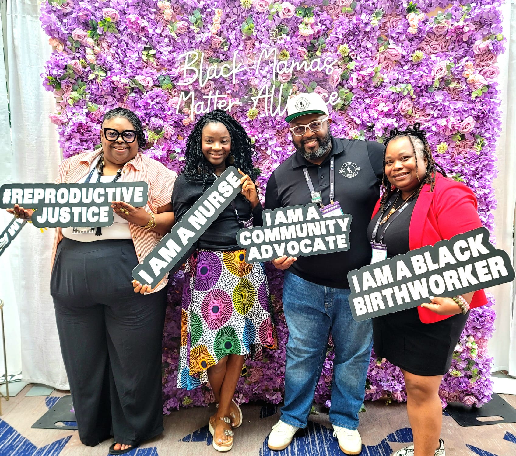 Four people stand in front of a purple flower backdrop holding signs supporting reproductive justice and community advocacy.