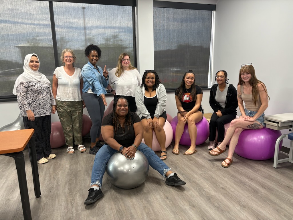Group of nine women in a room with large windows, some sitting on purple exercise balls, smiling.