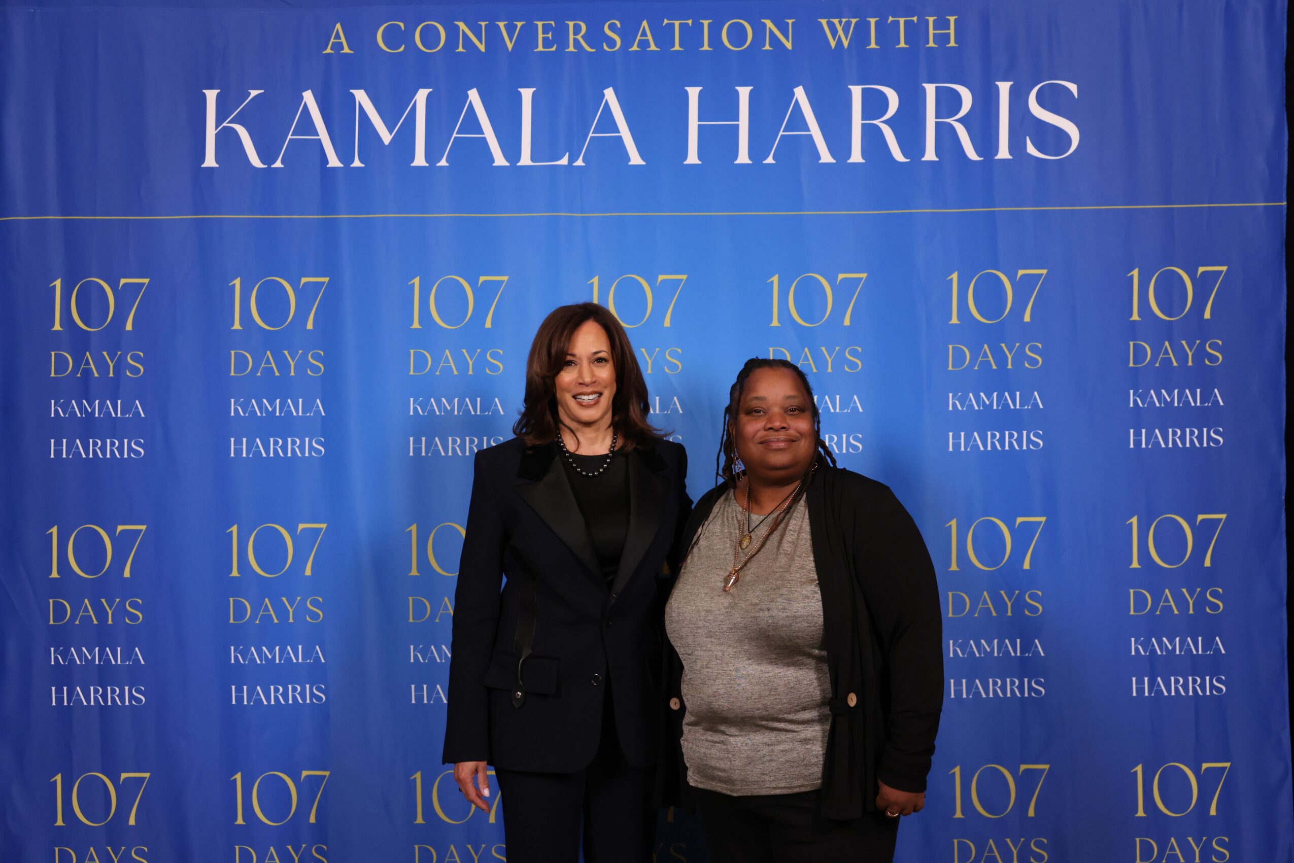 Two women standing in front of a blue backdrop with white text about Kamala Harris and 107 days.