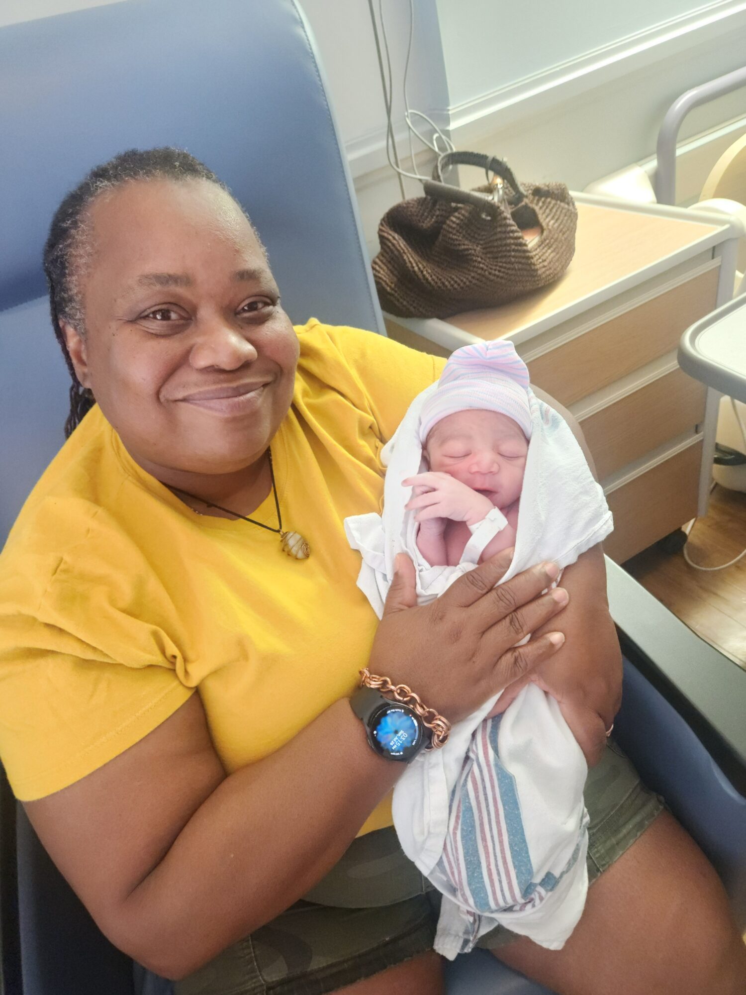 Person in yellow shirt holding a newborn baby wrapped in a white blanket, in a hospital room.