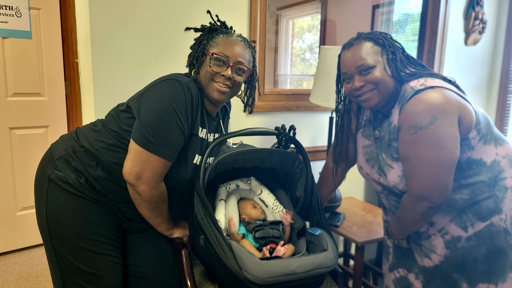 Two women smiling with a baby in a car seat indoors, near a window and a lamp, in a cozy room.