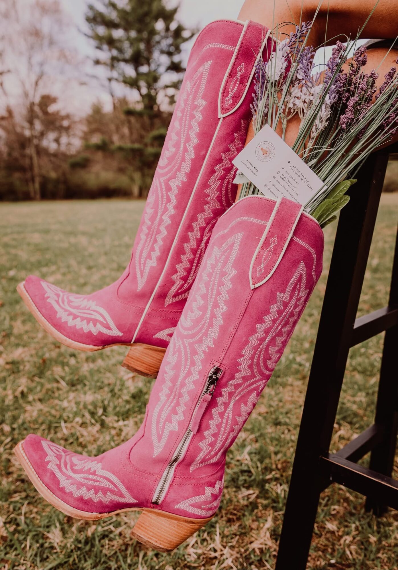 Pair of pink cowboy boots with decorative stitching, placed outdoors on grass, with a bouquet of lavender flowers inside one boot.