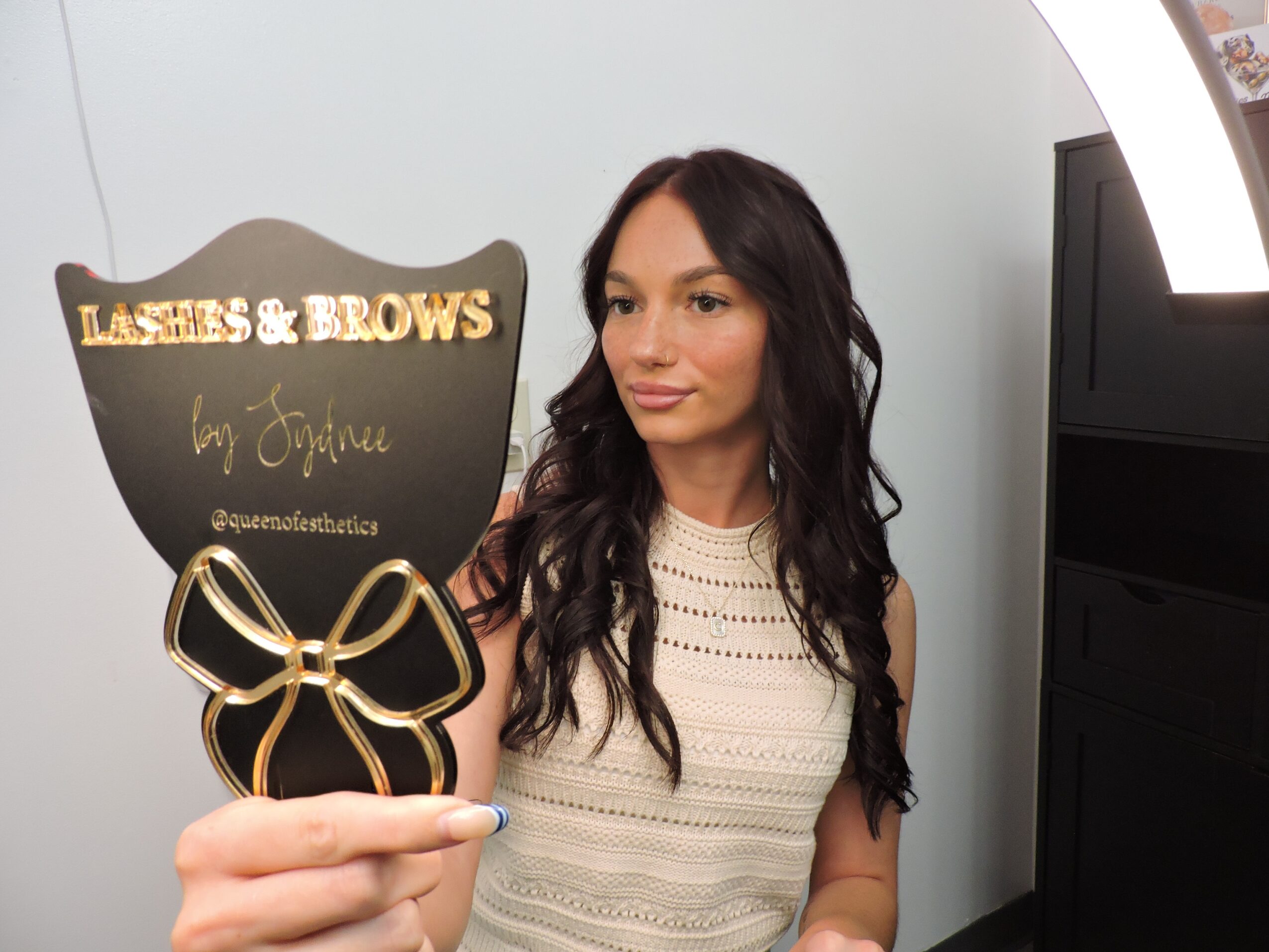 Woman with long dark hair holding a sign that says 'Lashes & Brows by Esthie' in a salon setting.