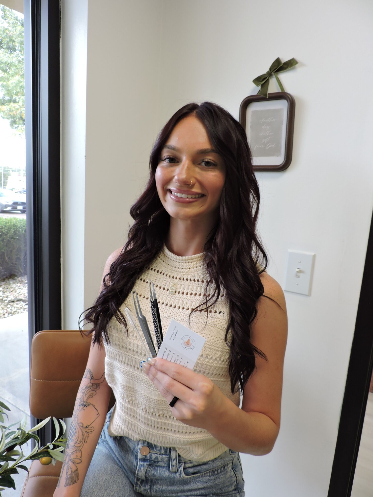 Young woman with long dark hair smiling, holding a phone, sitting indoors near a window with a plant.