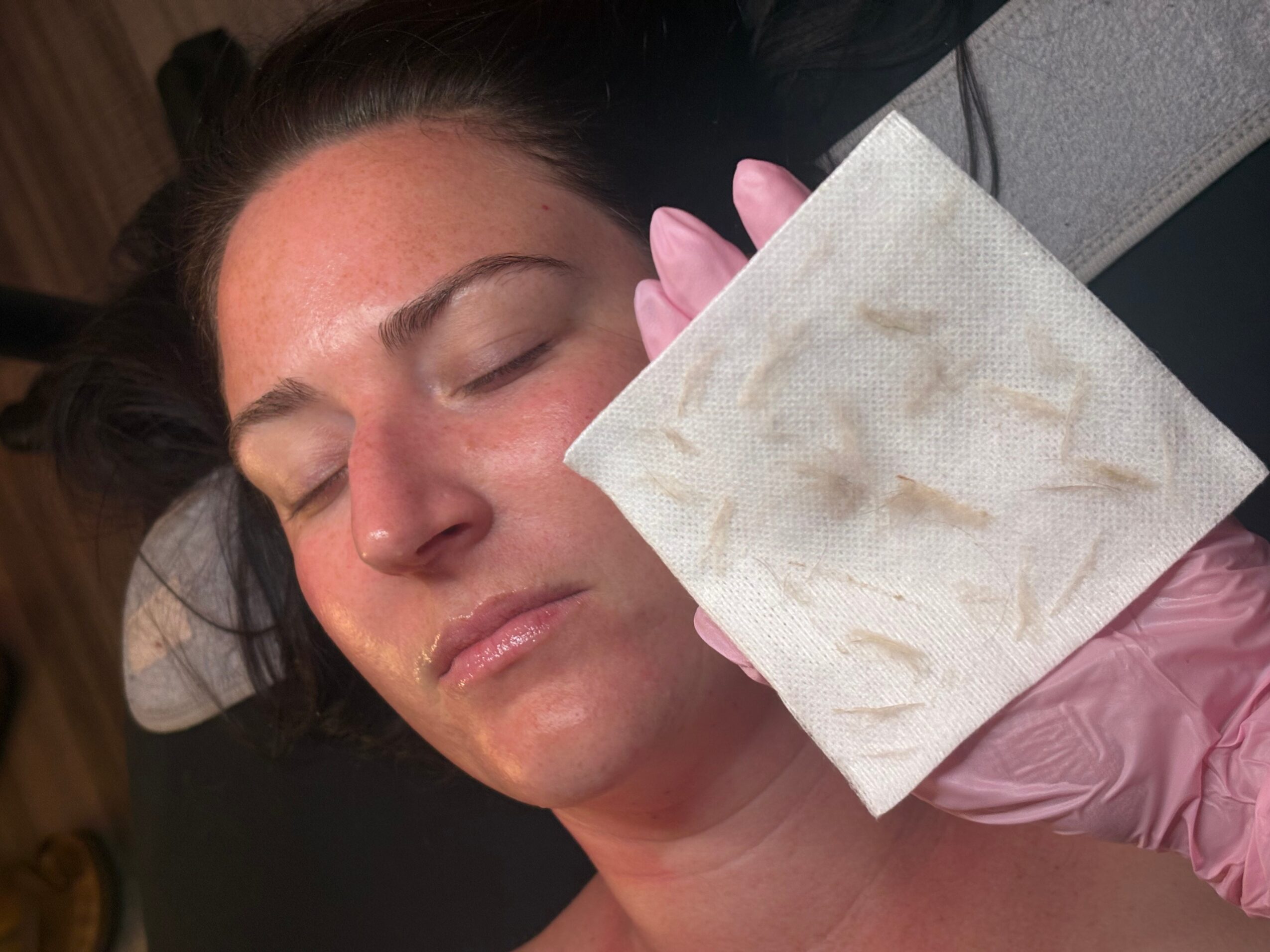 Woman with closed eyes receiving a facial treatment, holding a cotton pad, with a gloved hand nearby.
