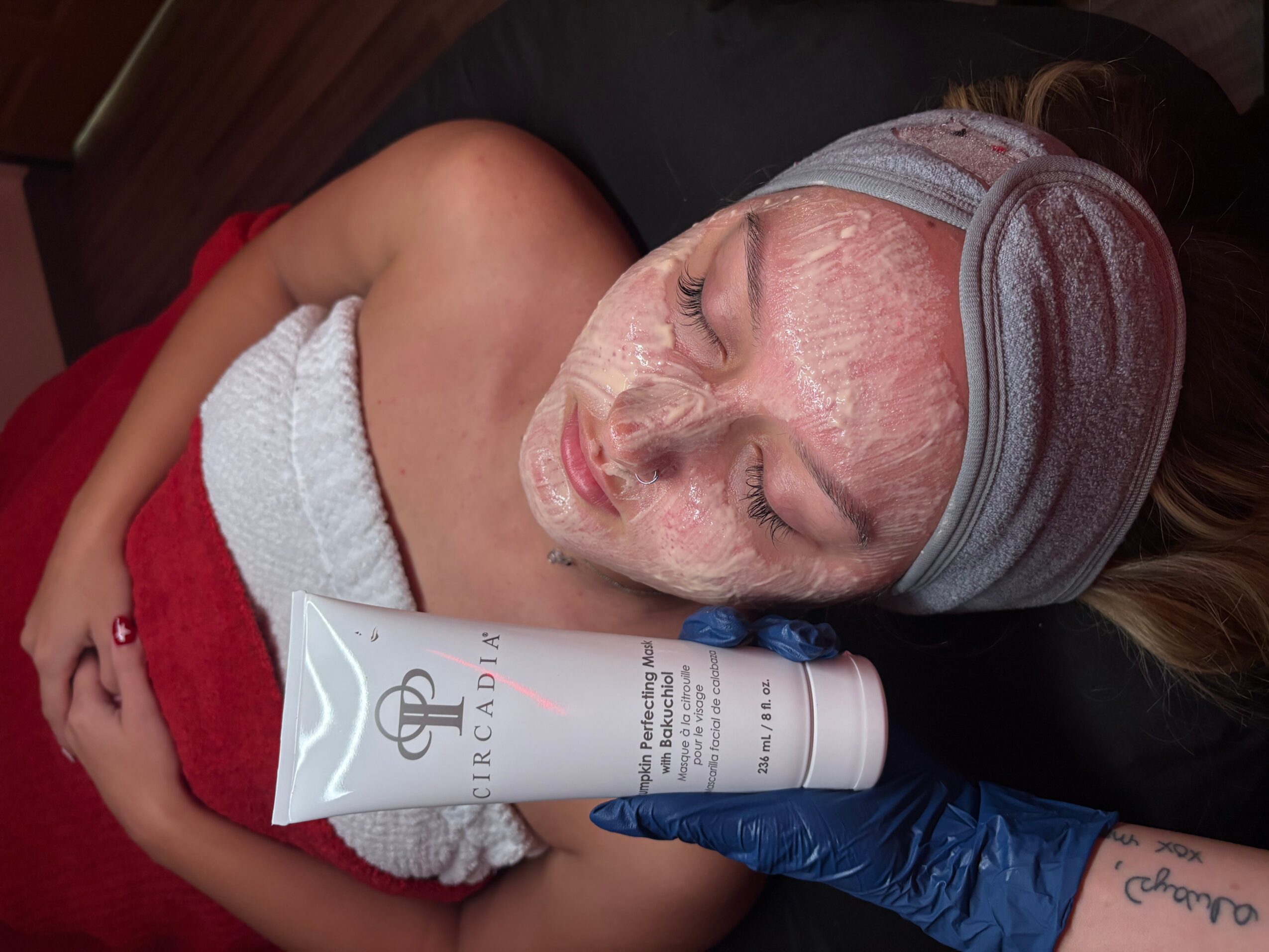 Person receiving facial treatment with a cream mask, lying down with eyes closed, head wrapped in a towel, and a professional applying product.