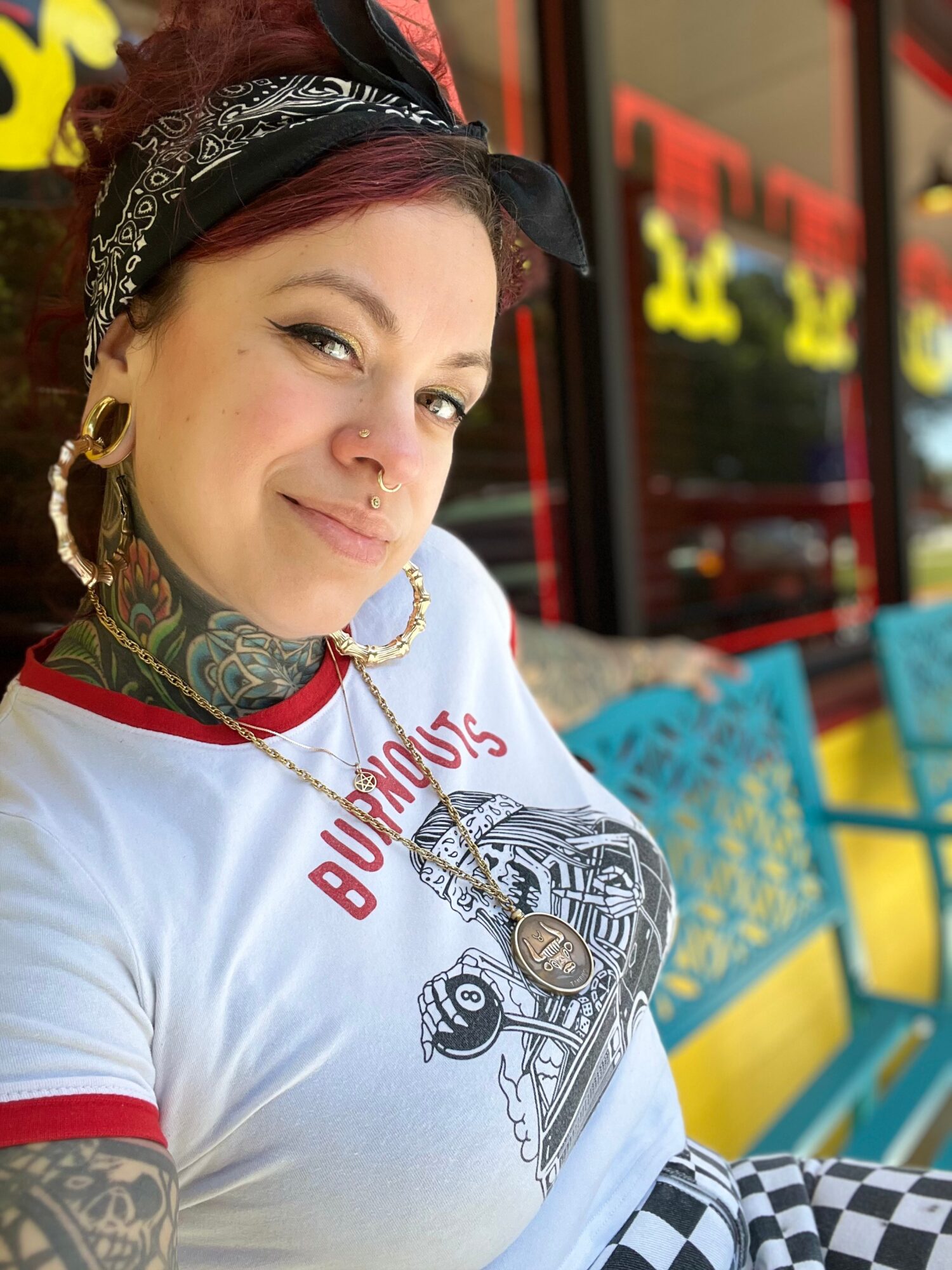 Woman with tattoos, piercings, and a bandana, smiling outside a colorful building with yellow and blue chairs.