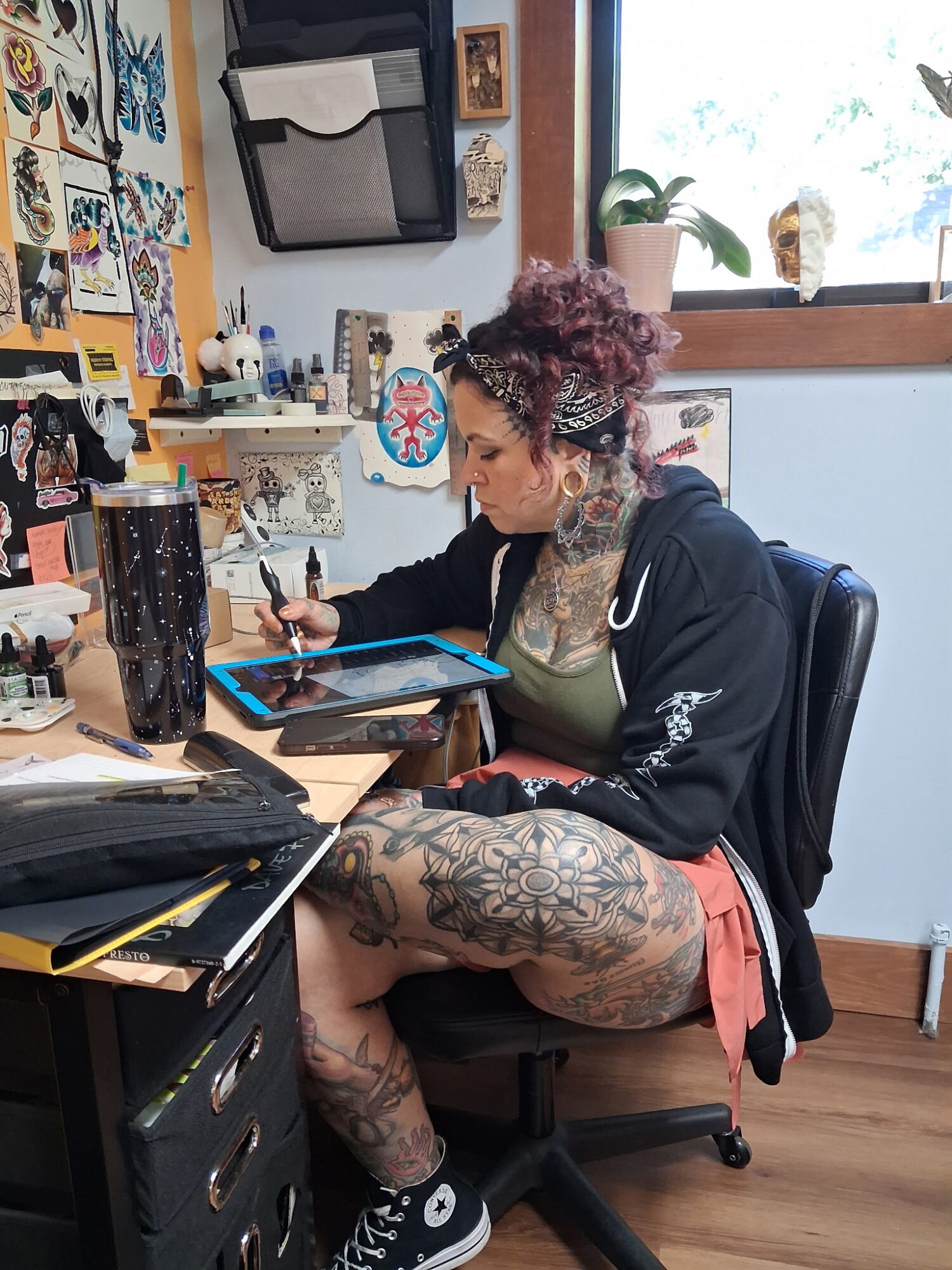Person with curly hair and tattoos sitting at a cluttered desk, using a tablet, in a room with artwork and a window.