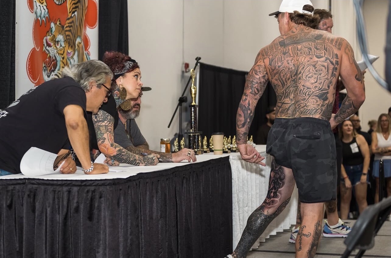Tattooed man with a cap stands at a table with two seated people, others watch in the background.