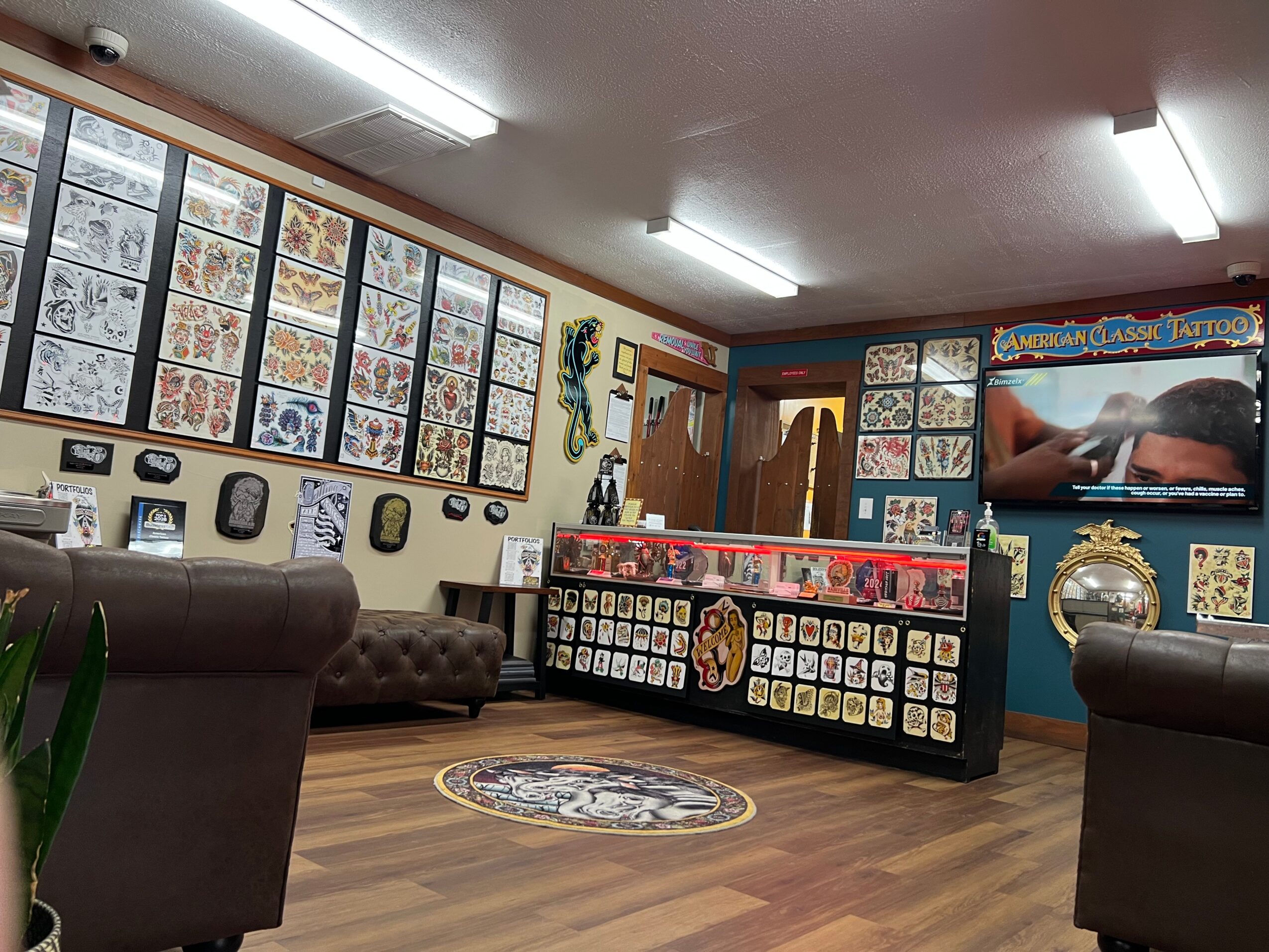 Tattoo shop interior with framed tattoo art, seating, display counter, and a television screen, wooden floor, and bright lighting.