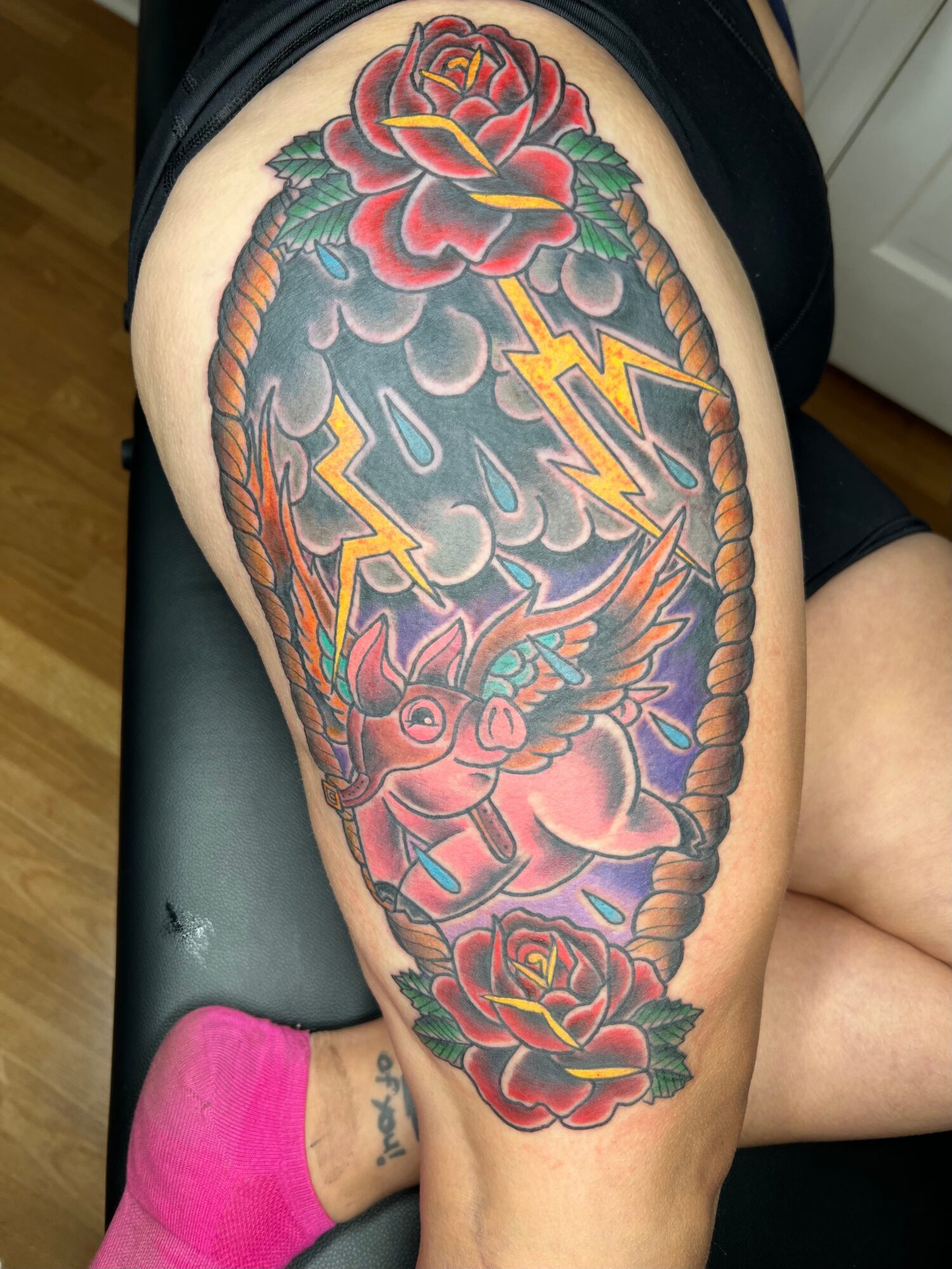 Tattoo of a pink winged creature with lightning bolts, surrounded by red roses and green leaves on upper thigh.