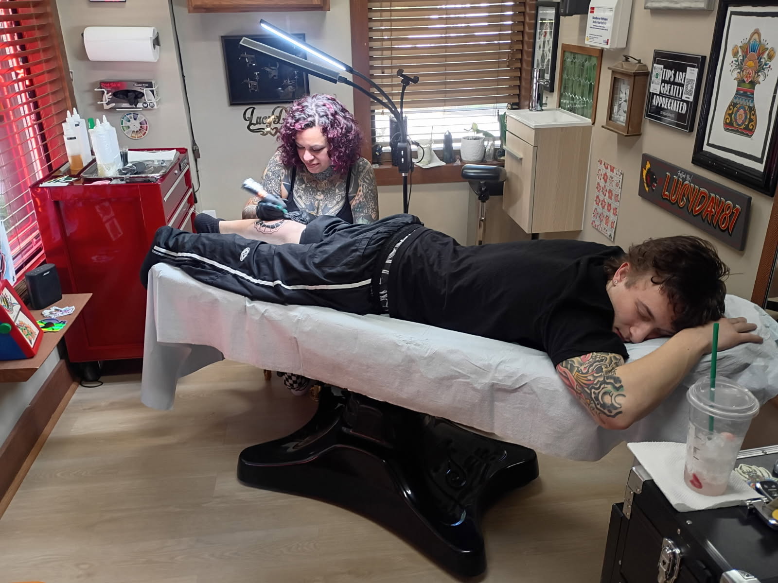 Person lying face down on a tattoo table in a tattoo studio, with artist working nearby, surrounded by framed artwork and equipment.