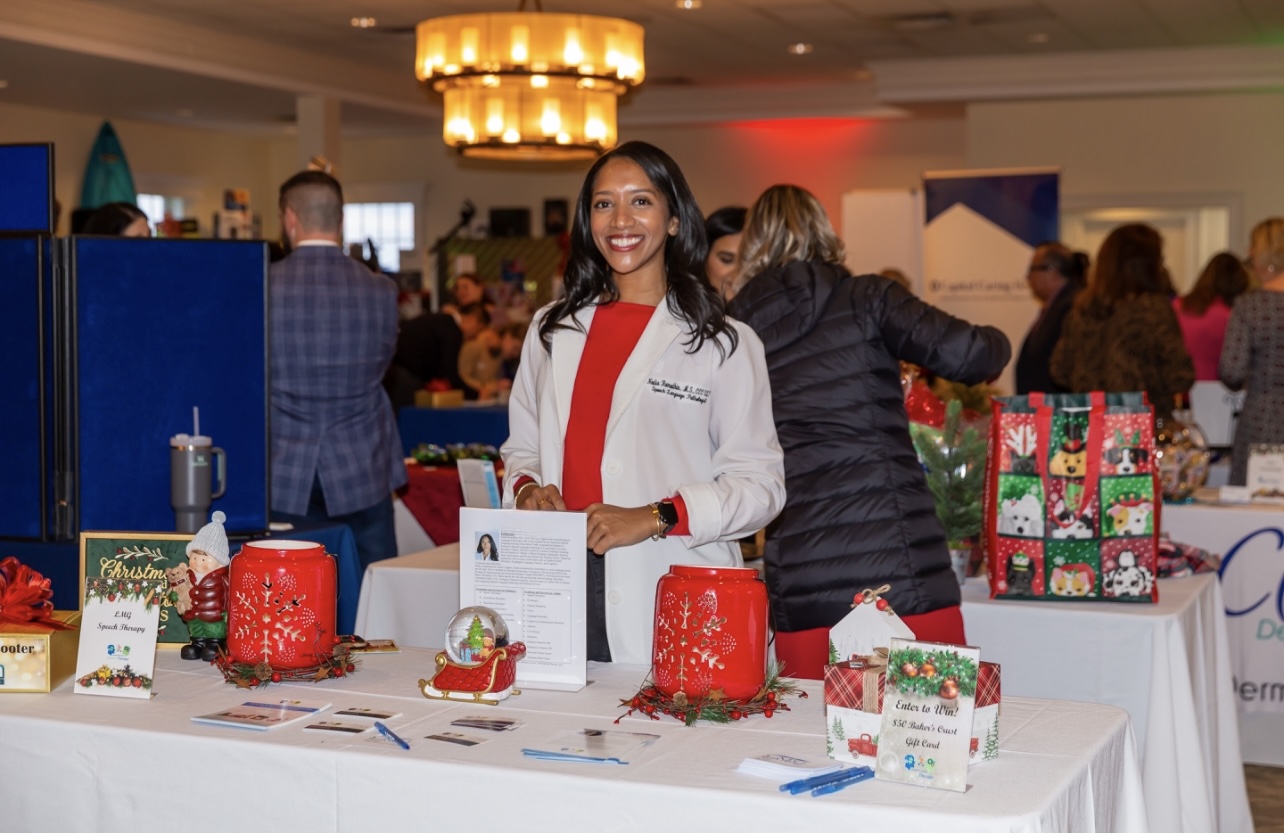 Smiling woman in white coat standing behind a table with Christmas gifts and decorations at an indoor event.