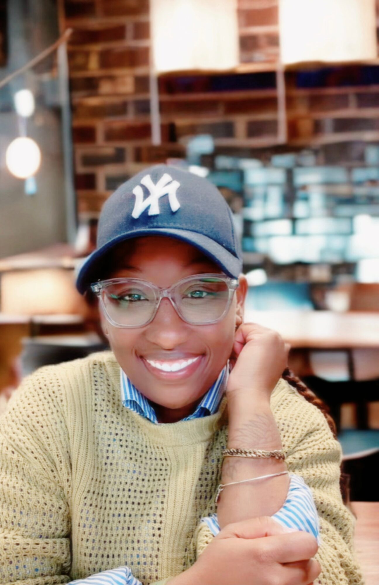 Smiling person wearing glasses, a New York Yankees cap, and a yellow sweater in a cozy indoor setting.