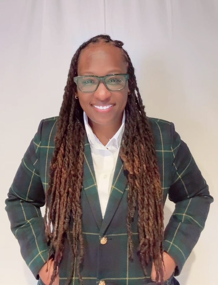 Woman with long dreadlocks, glasses, smiling, wearing a dark plaid blazer and white shirt, standing with hands on hips.
