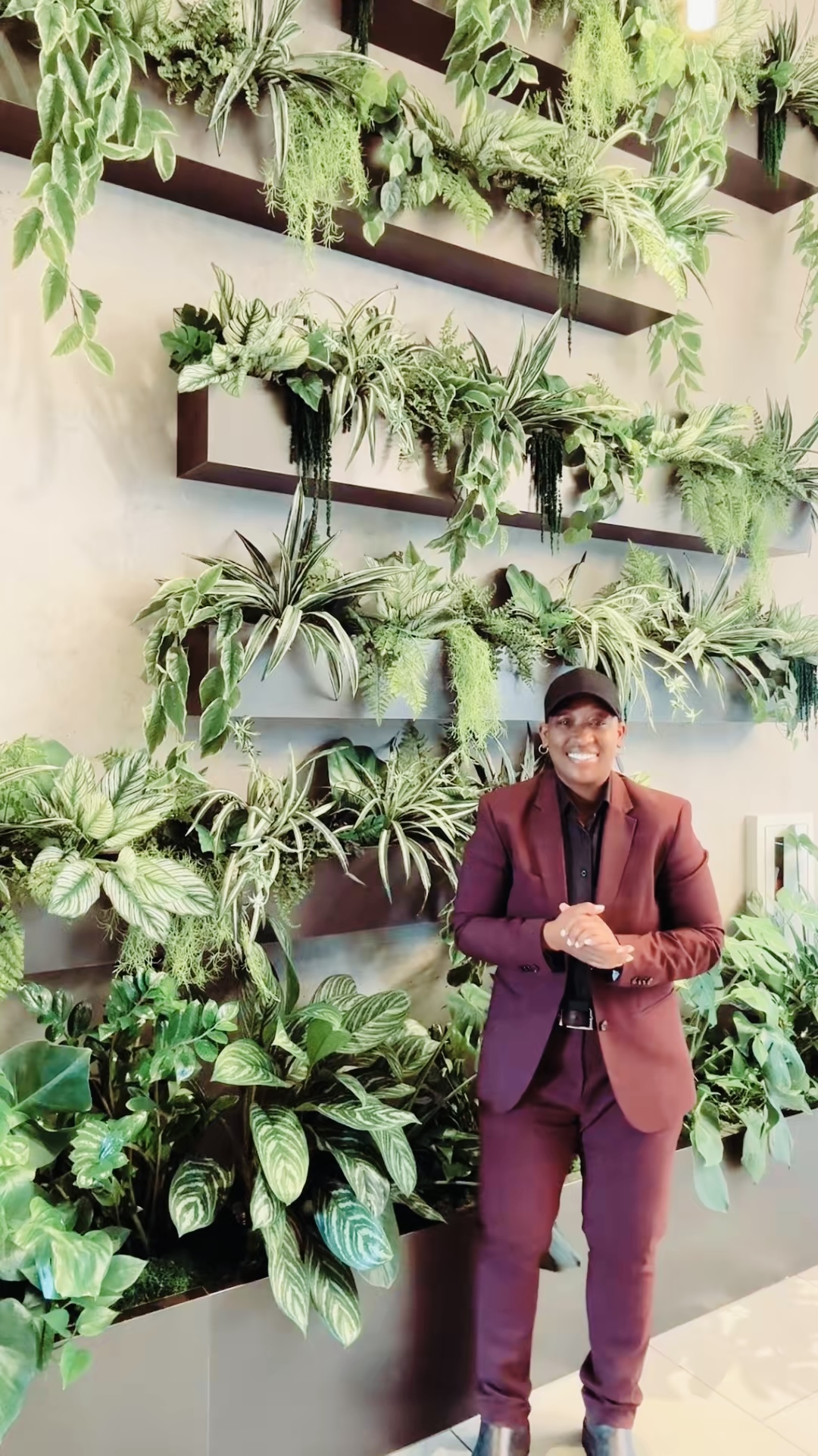 Person in maroon suit and glasses standing in front of a wall with green plants and shelves.