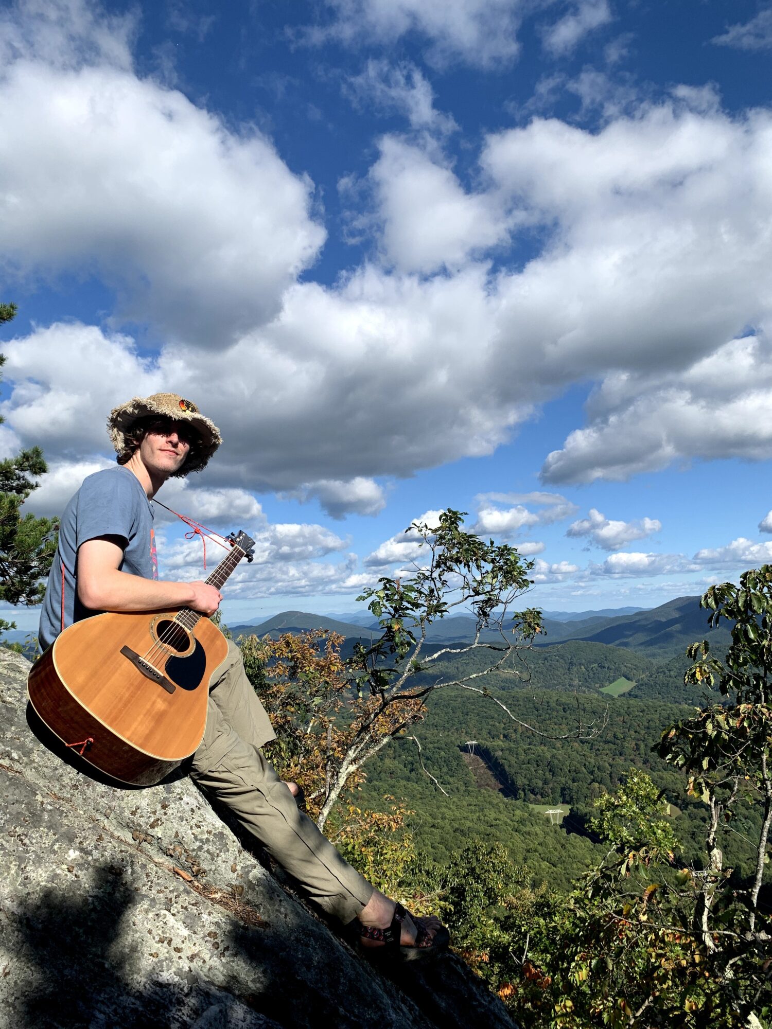 Person sitting on a rock holding a guitar, overlooking a green landscape with trees and mountains under a partly cloudy sky.