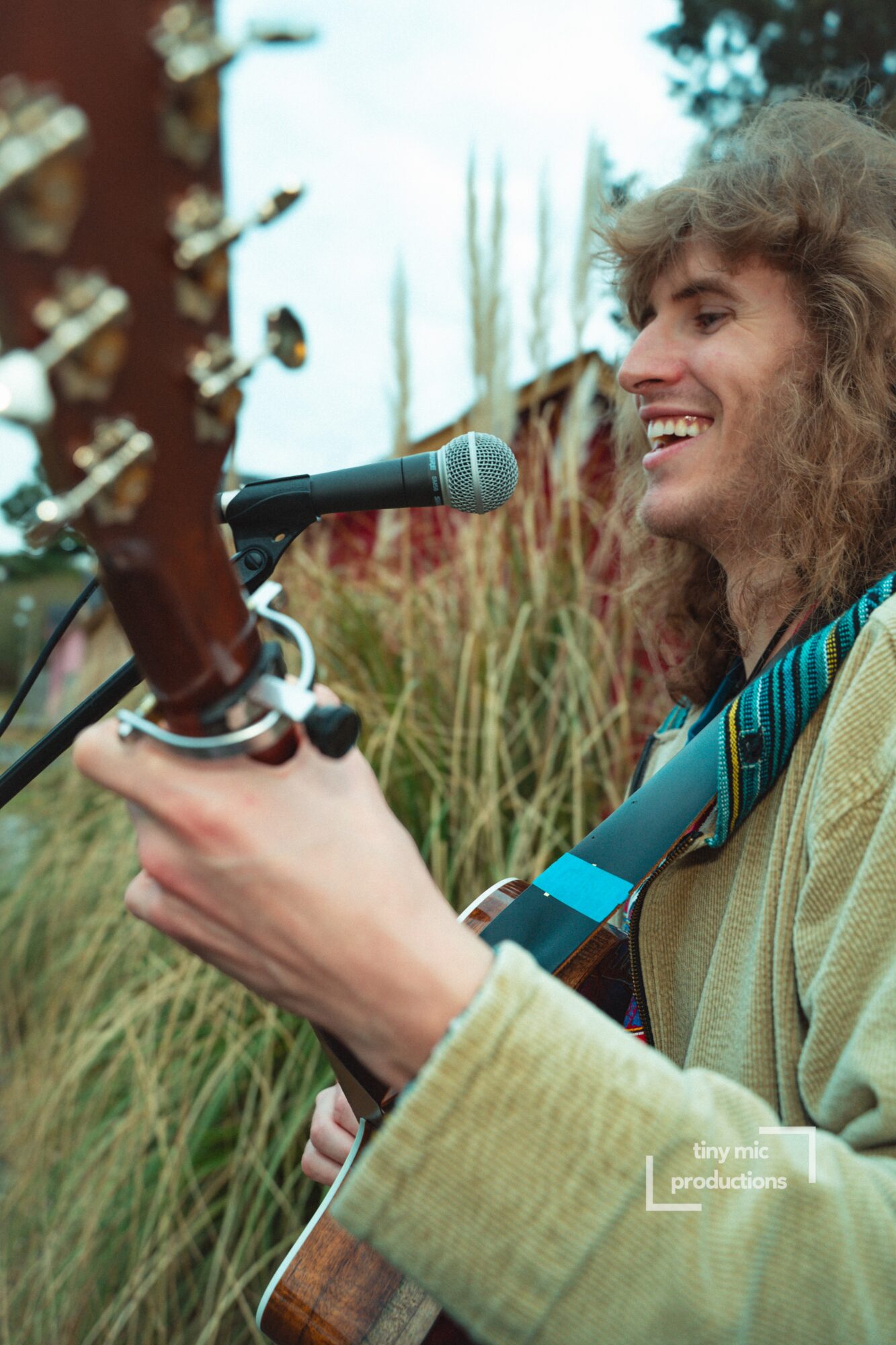 Person with curly hair smiling, holding a guitar and singing into a microphone outdoors, surrounded by tall grass and trees.