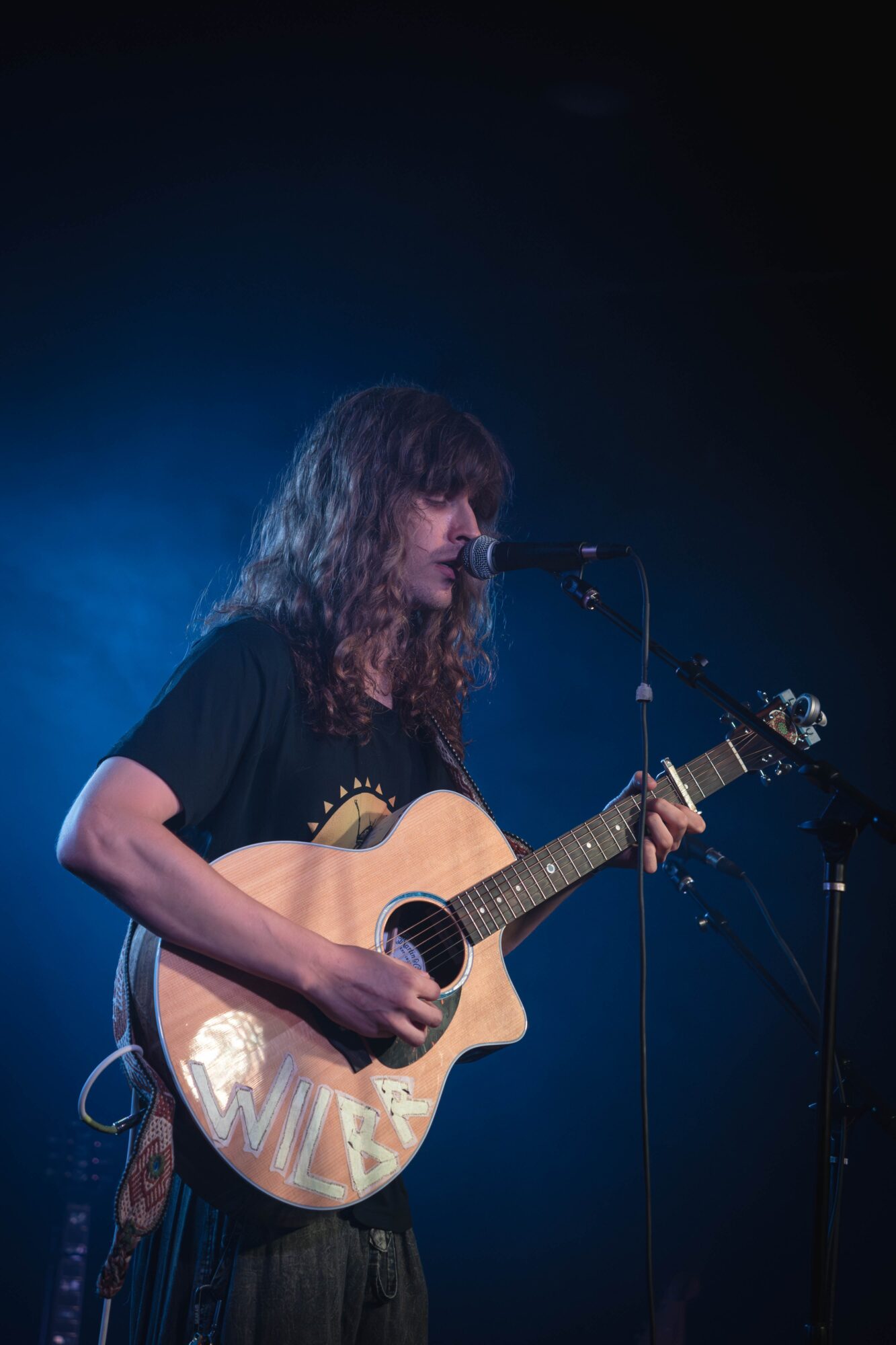 Person with long hair playing an acoustic guitar on stage with blue lighting, microphone nearby.