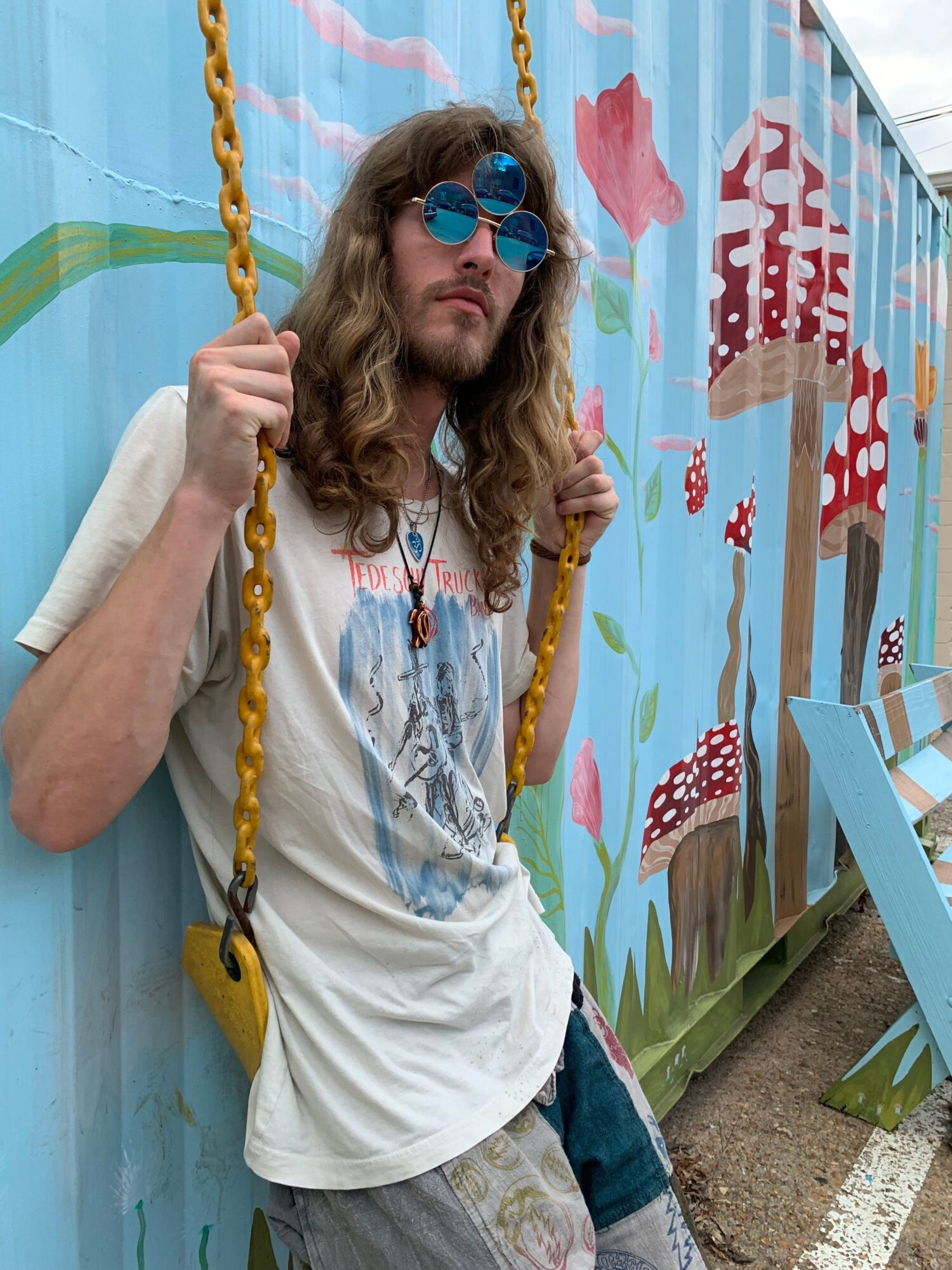 Person with long hair, sunglasses, and a white shirt, holding yellow swings, standing next to a colorful fence.