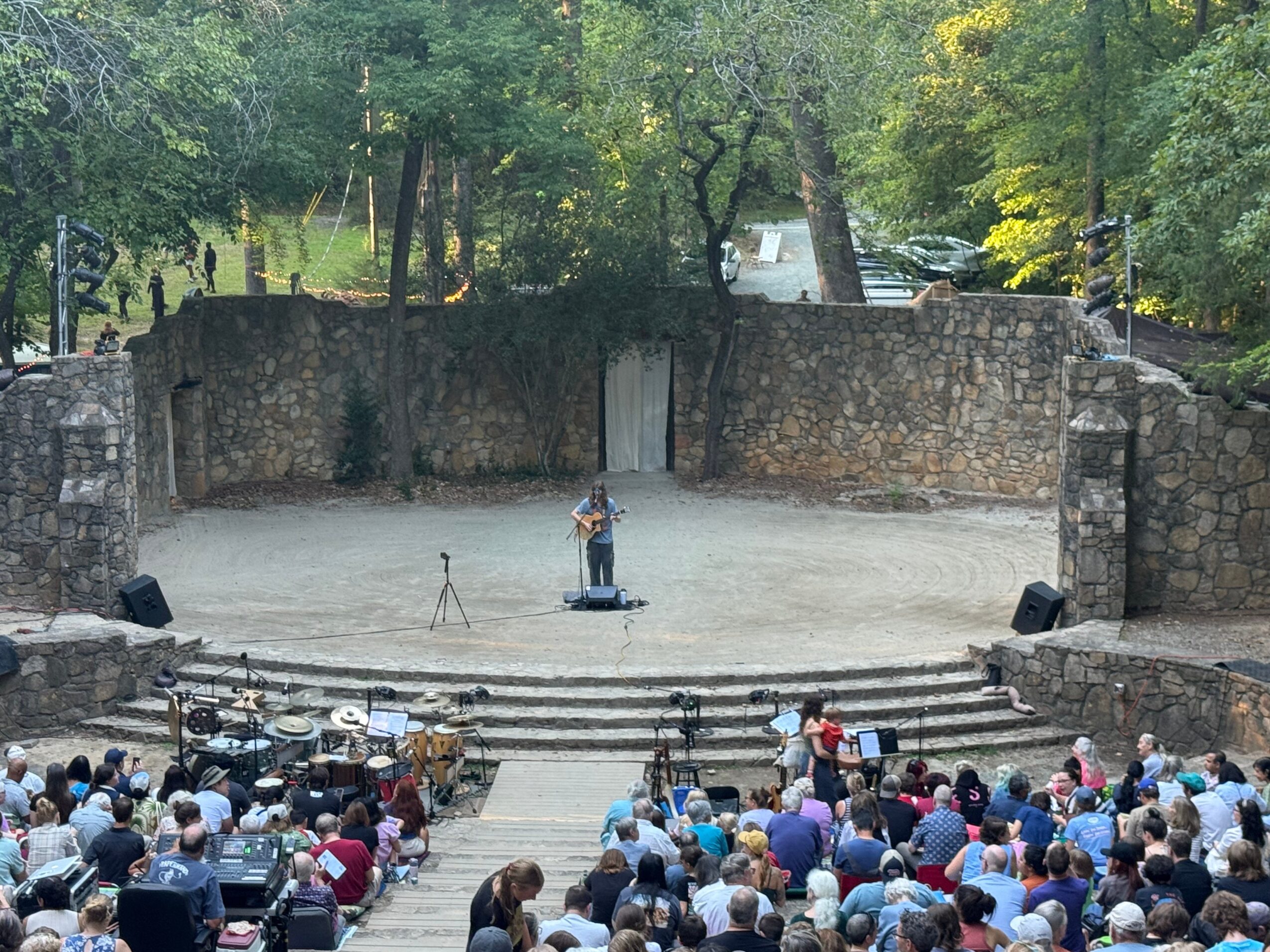 Open-air outdoor theater with a stone stage, audience seated on steps, surrounded by trees, performer on stage with equipment.