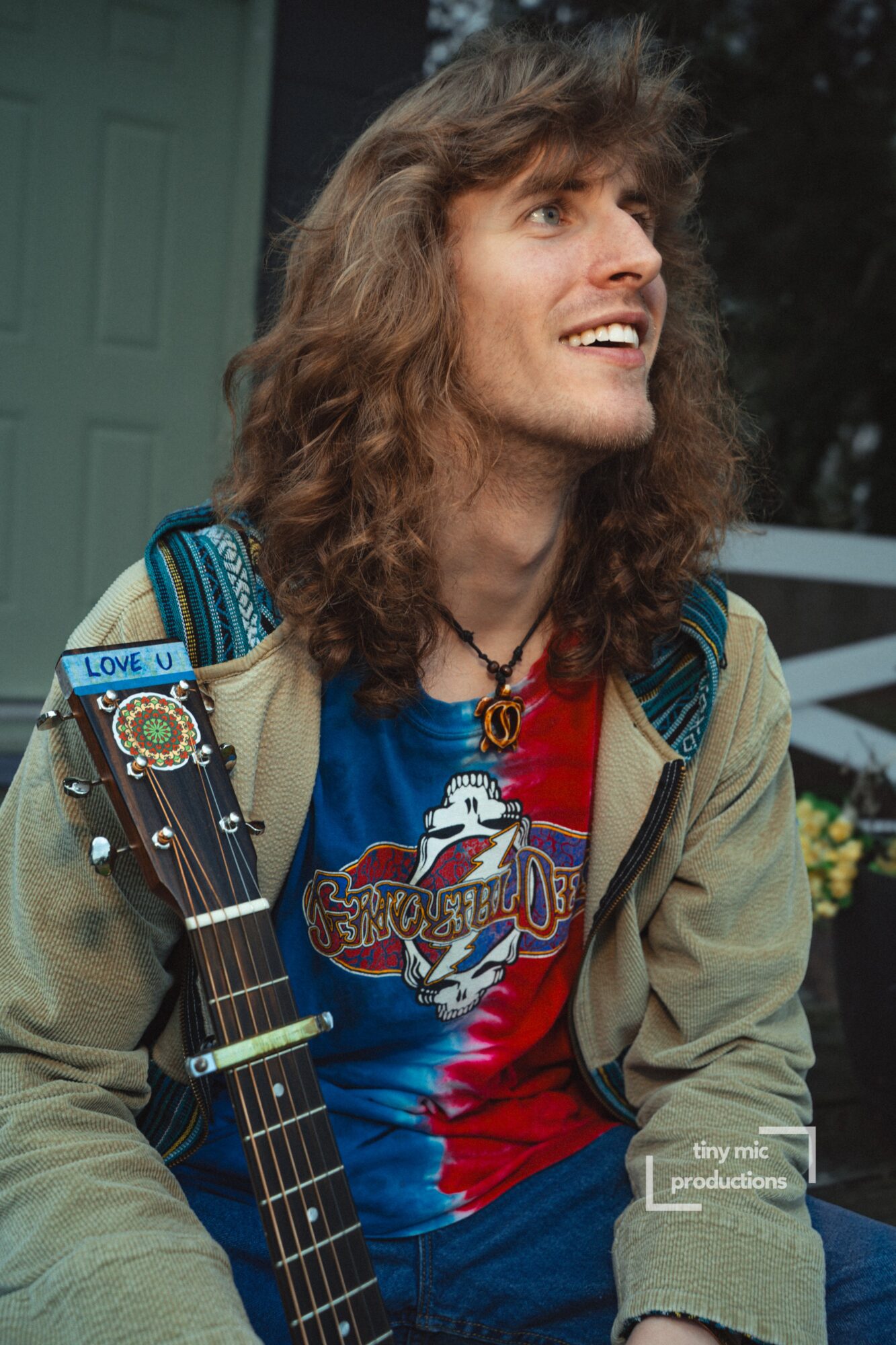 Young man with long, curly hair smiling, holding a guitar, wearing a colorful T-shirt and jacket outdoors.