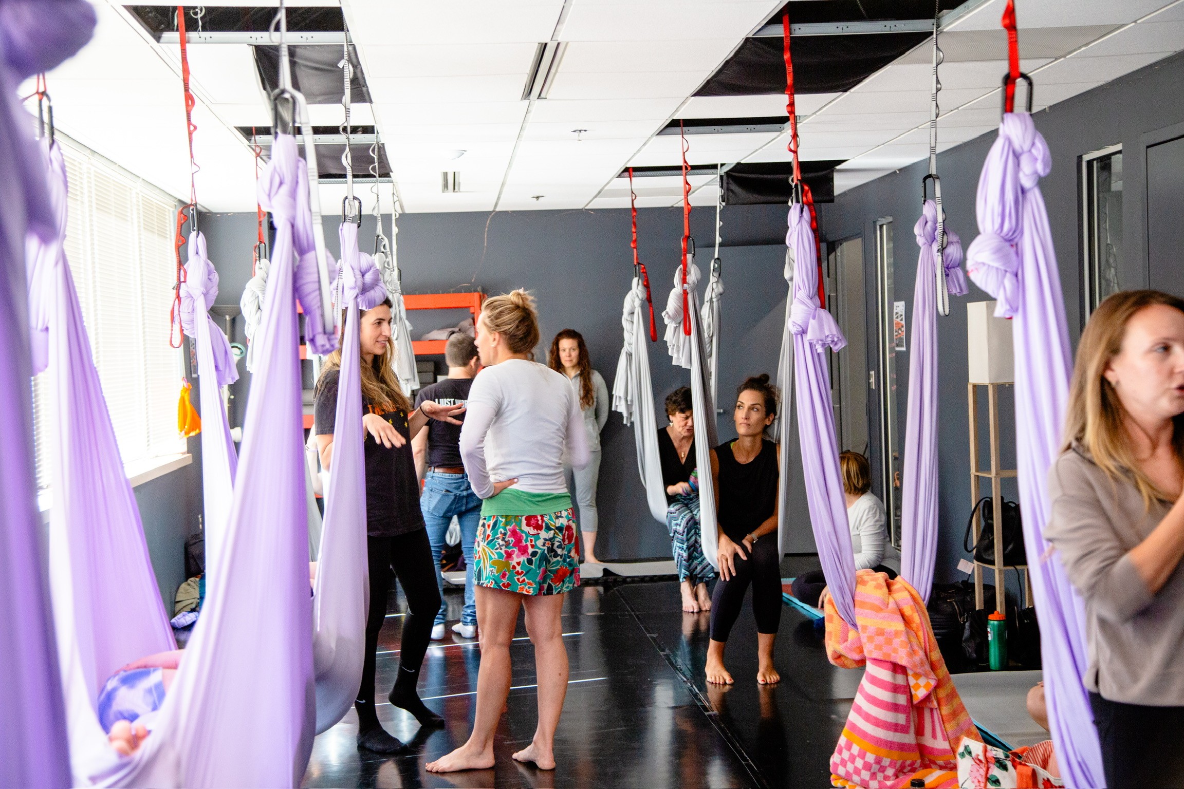 Group of people, mostly women and children, in a room with purple fabric swings hanging from the ceiling, some sitting and some standing.