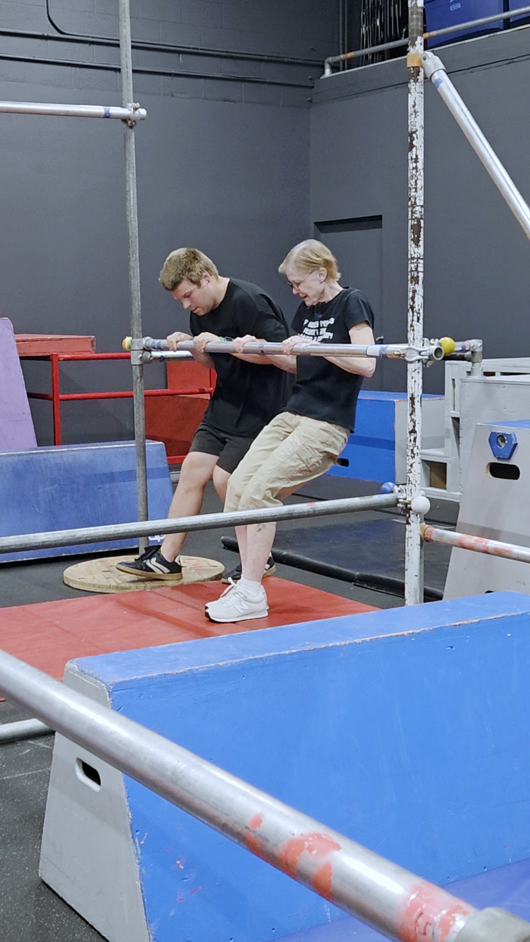 Two young men are on a trampoline surrounded by metal safety bars indoors.