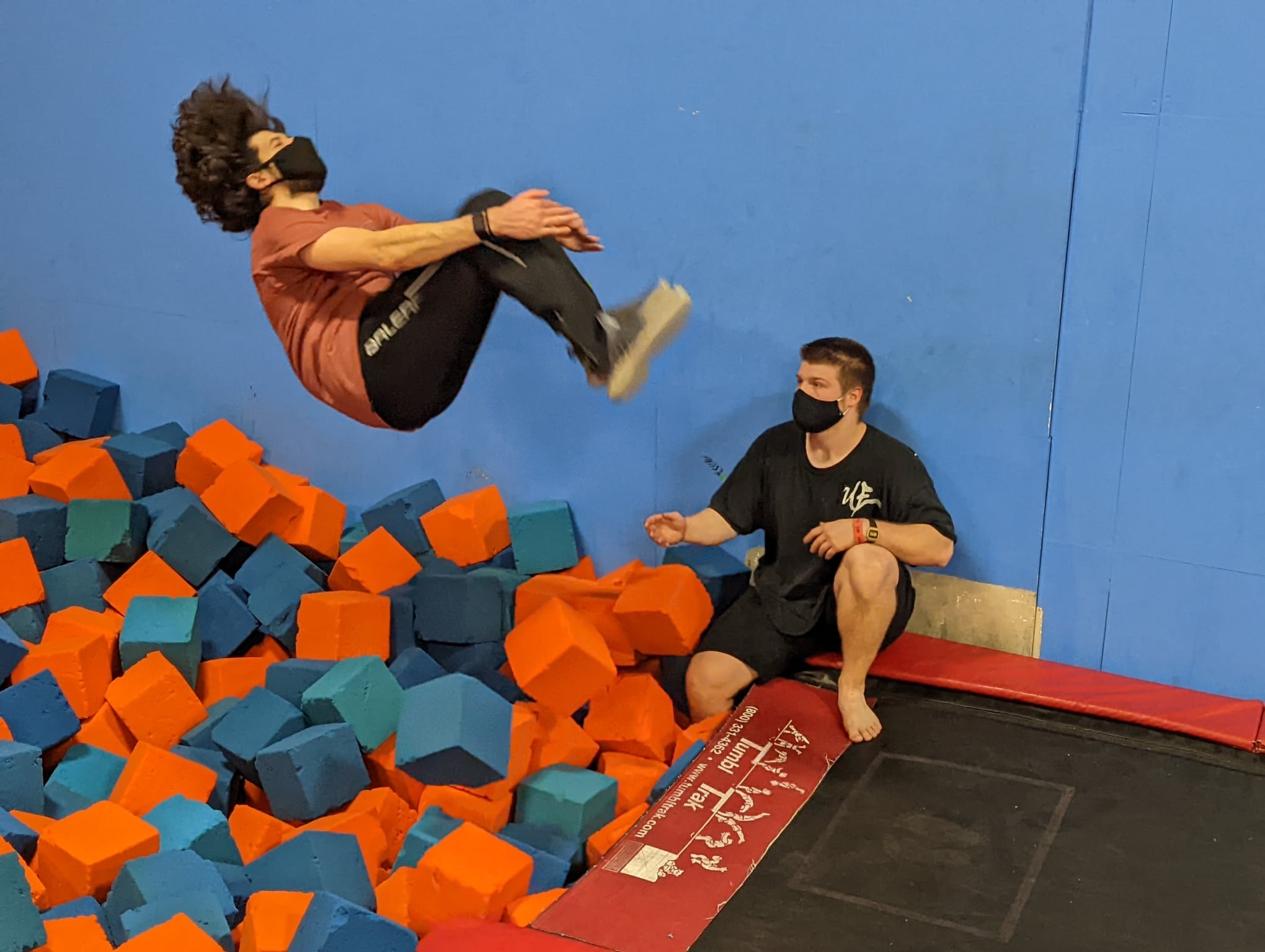 Person jumping into foam pit while another person sits nearby against a blue wall, both wearing masks.
