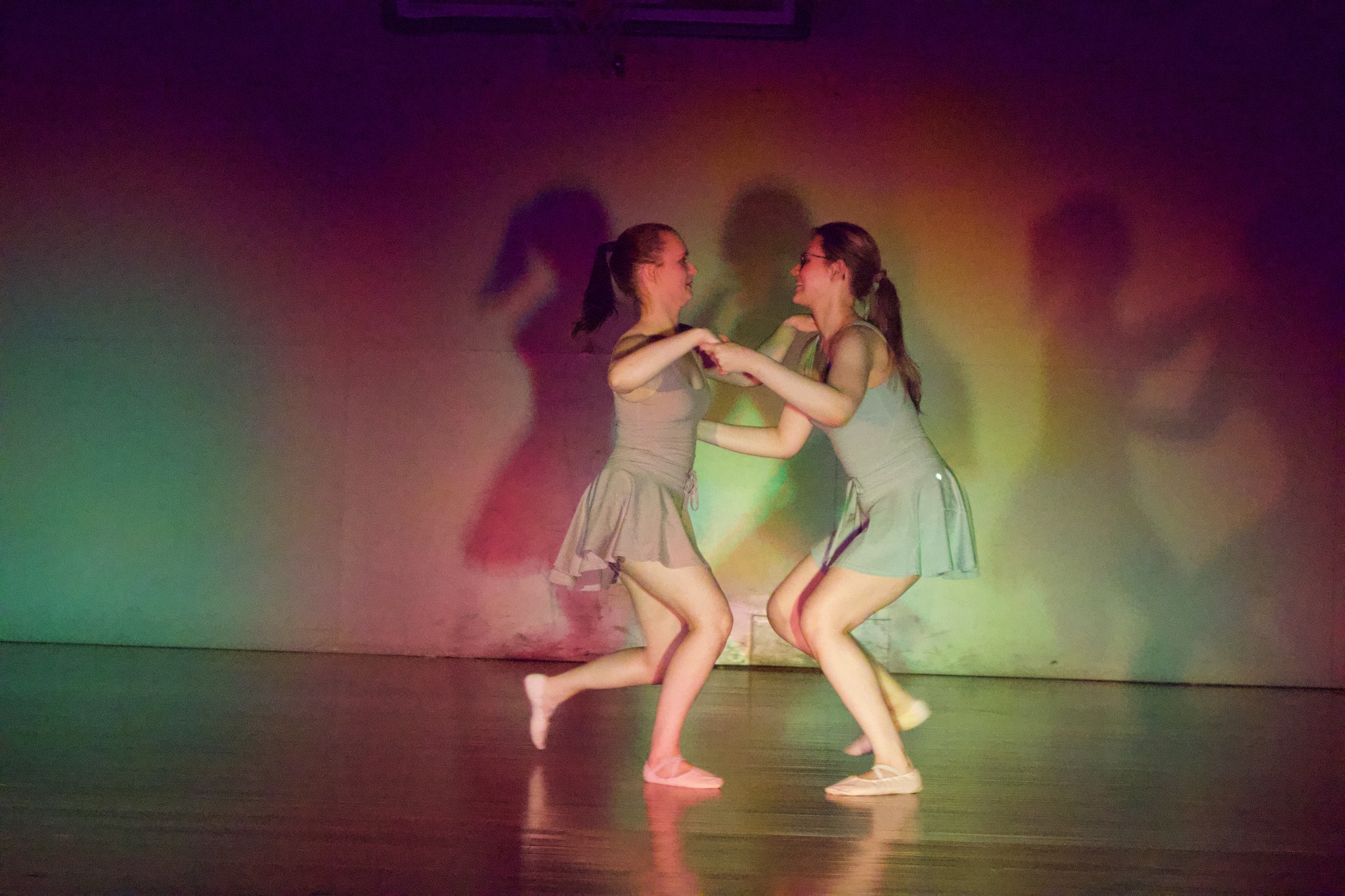 Two young girls dance together on stage, holding hands, with colorful lighting and shadows on the background wall.
