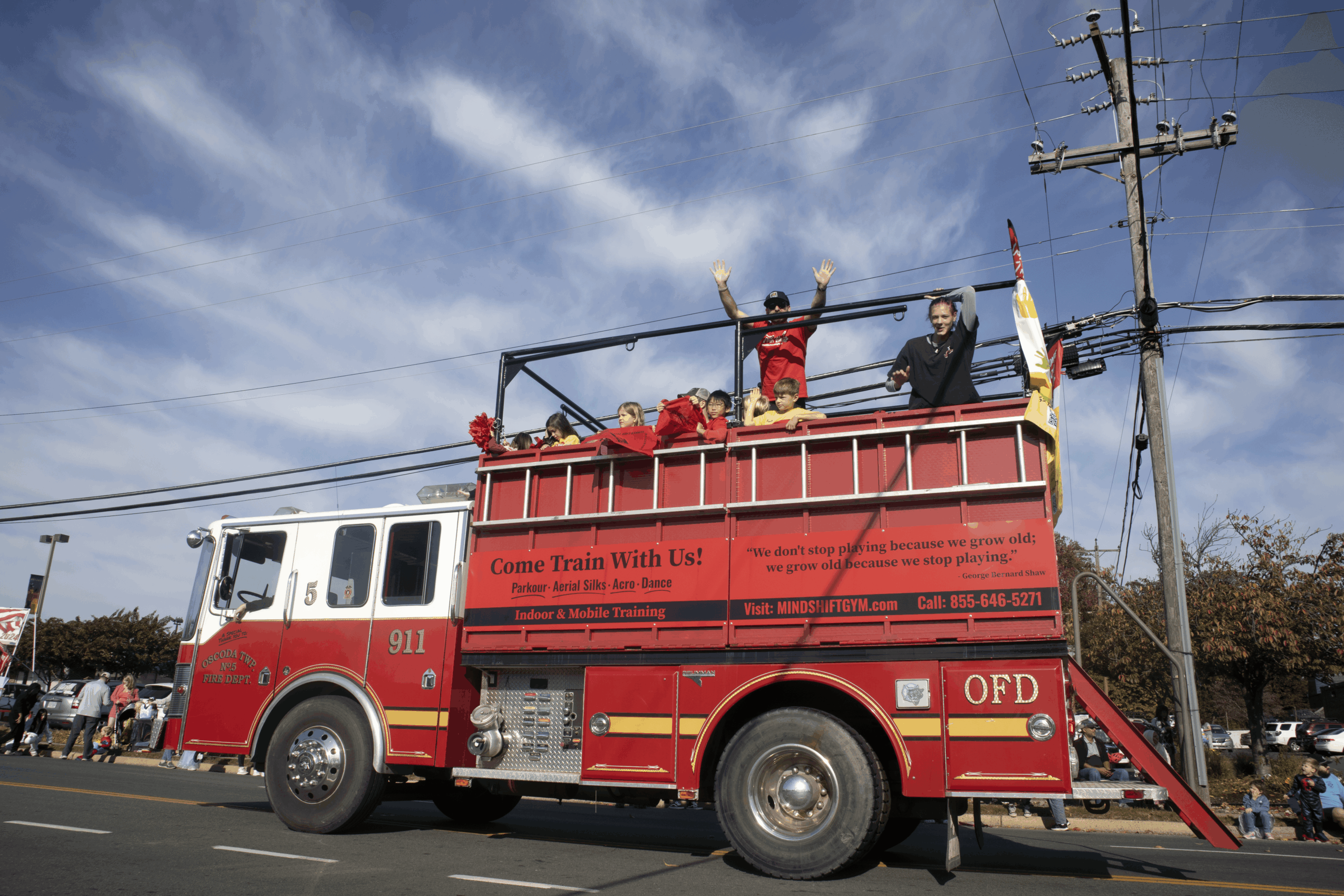 Firefighters on a red fire truck's upper platform waving, with a blue sky and power lines in the background.