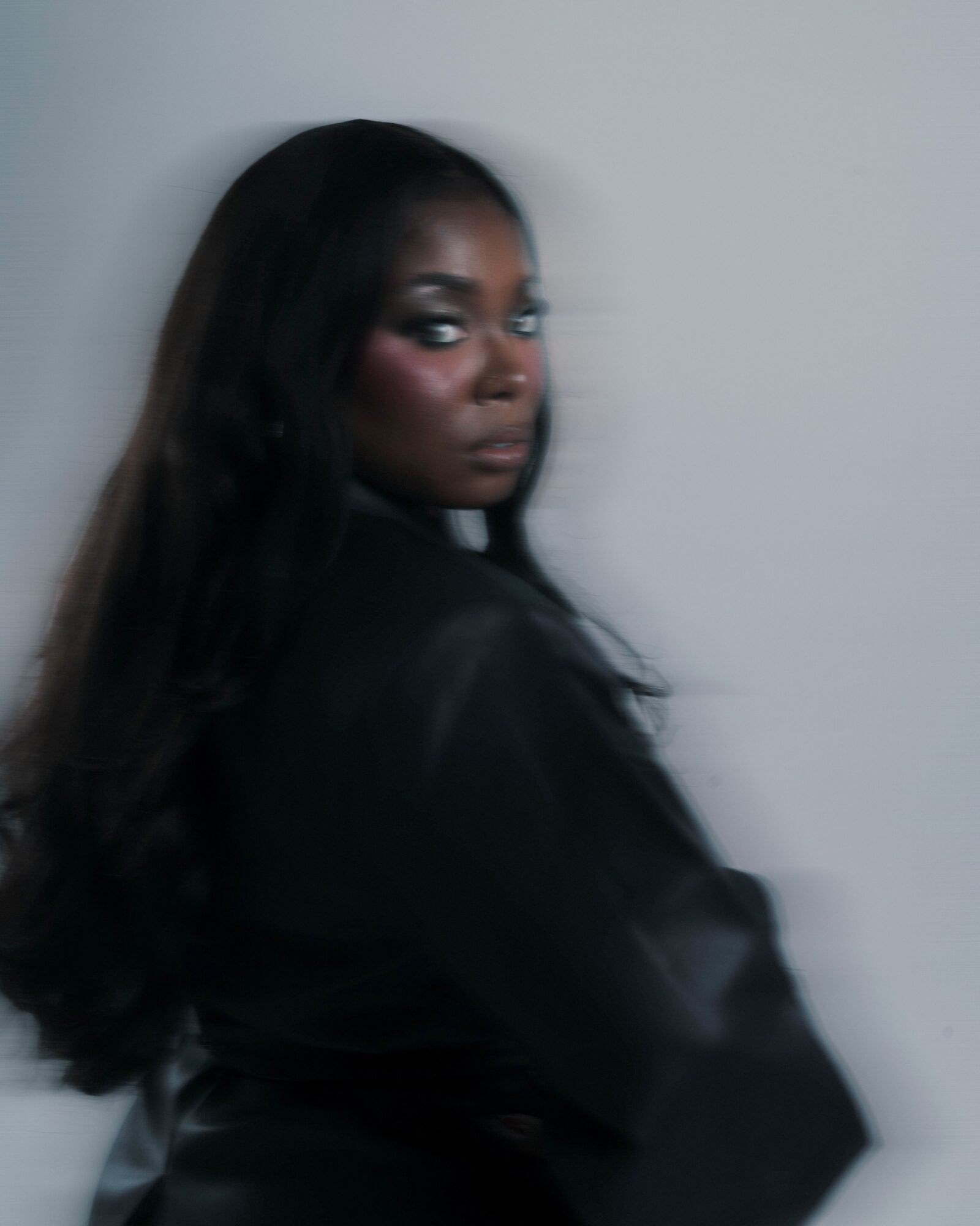 Woman with long dark hair looking over shoulder, dark makeup, black clothing, against a plain light background.