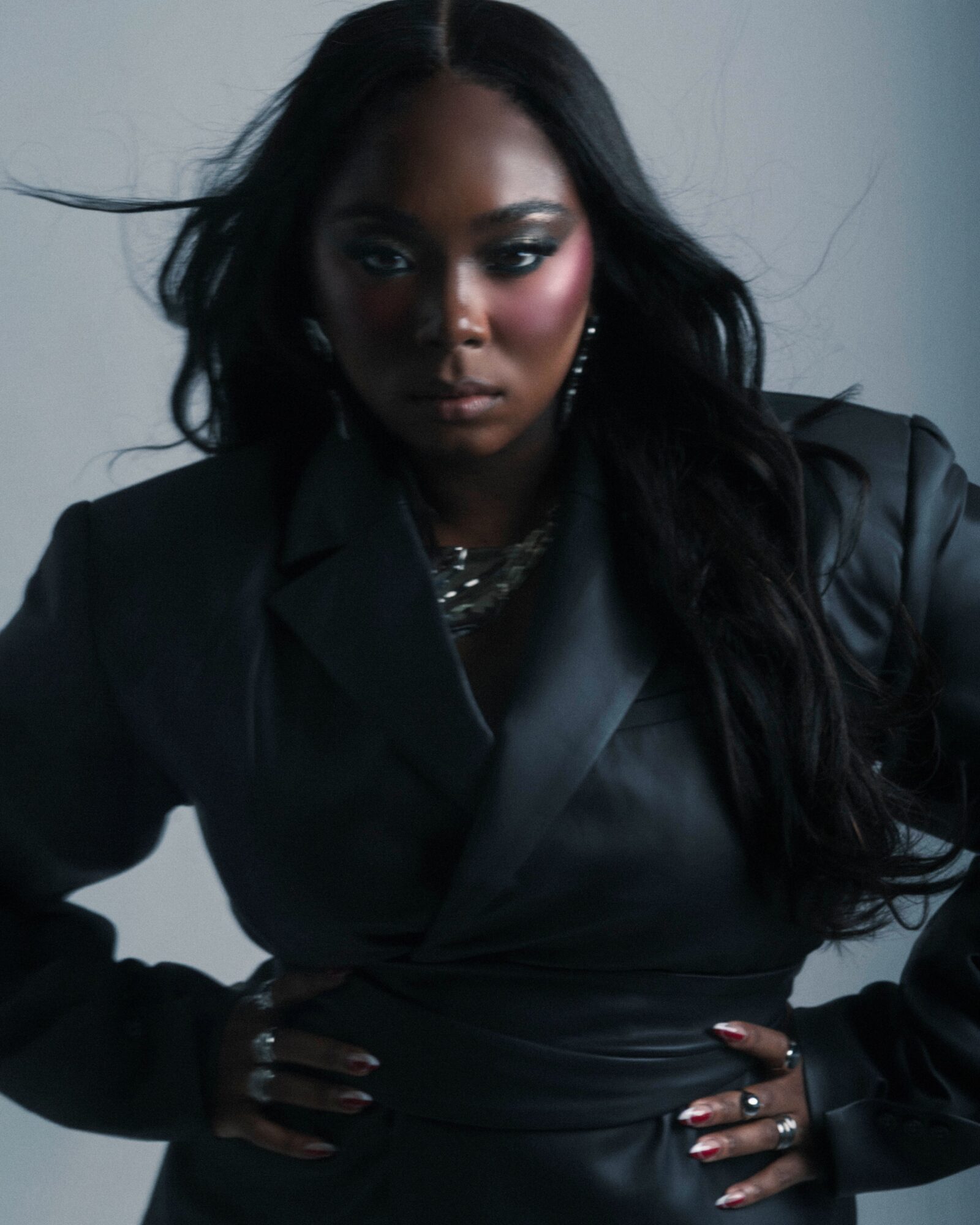 Woman with long dark hair wearing a black leather jacket, posing with hands on hips, against a plain background.