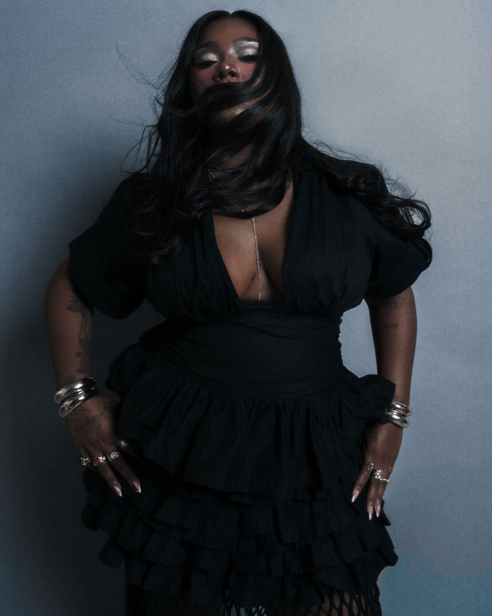 Woman with long dark hair wearing sunglasses, black dress, and jewelry, standing against a plain background.