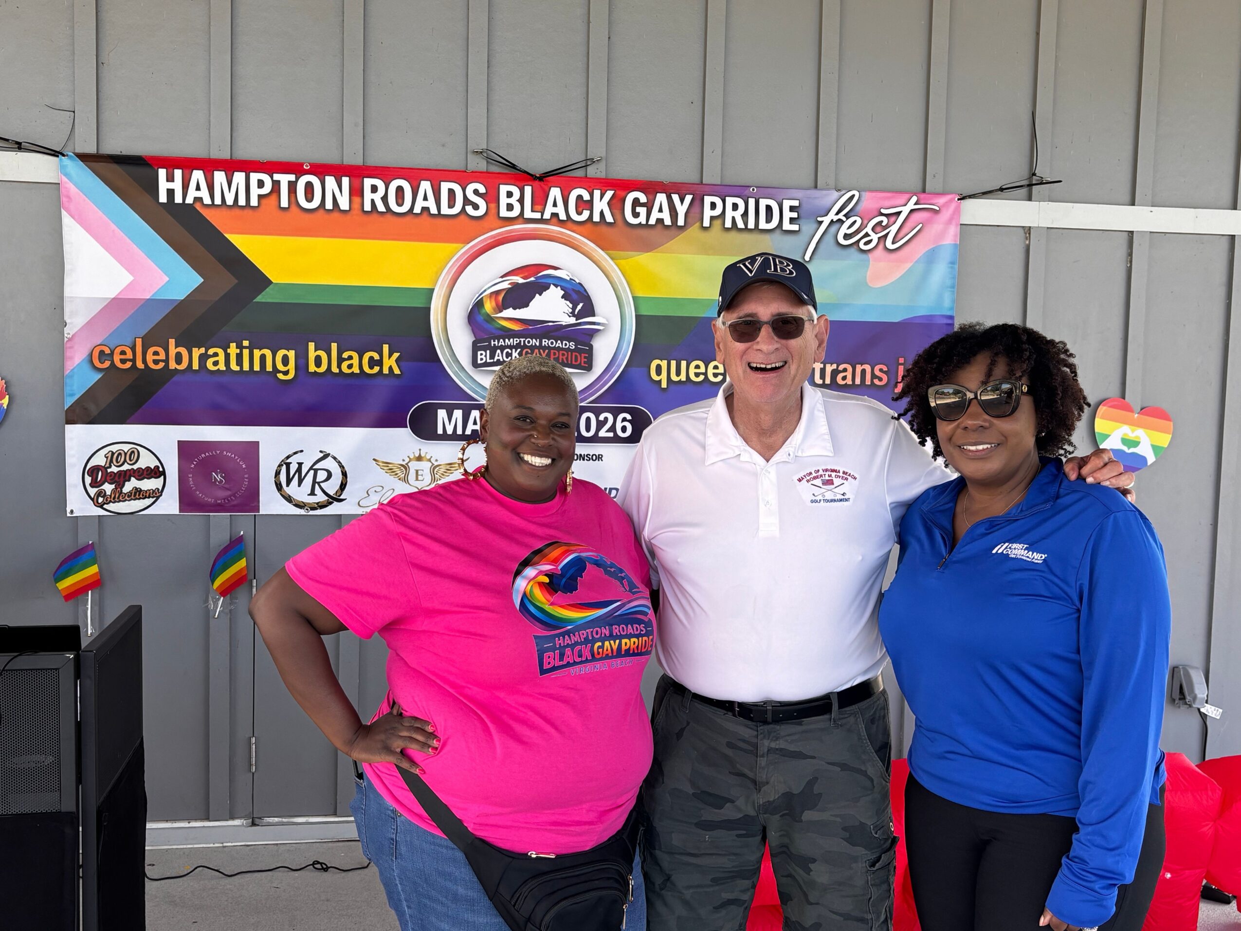 Two people standing in front of a banner, one wearing a pink shirt and the other in white, both holding rainbow flags.