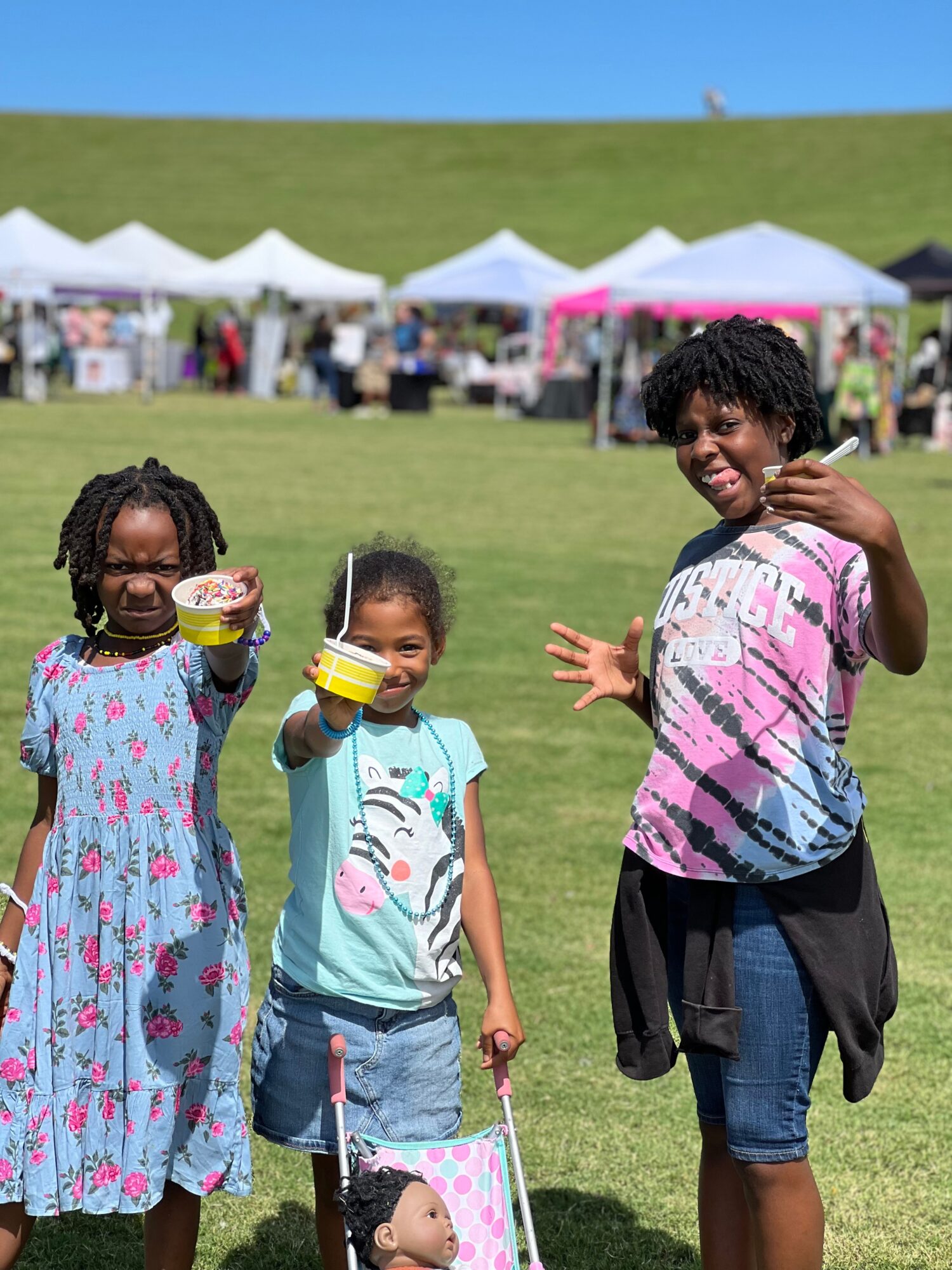 Three children smiling and holding food and a bag outdoors with tents and a grassy hill in the background.