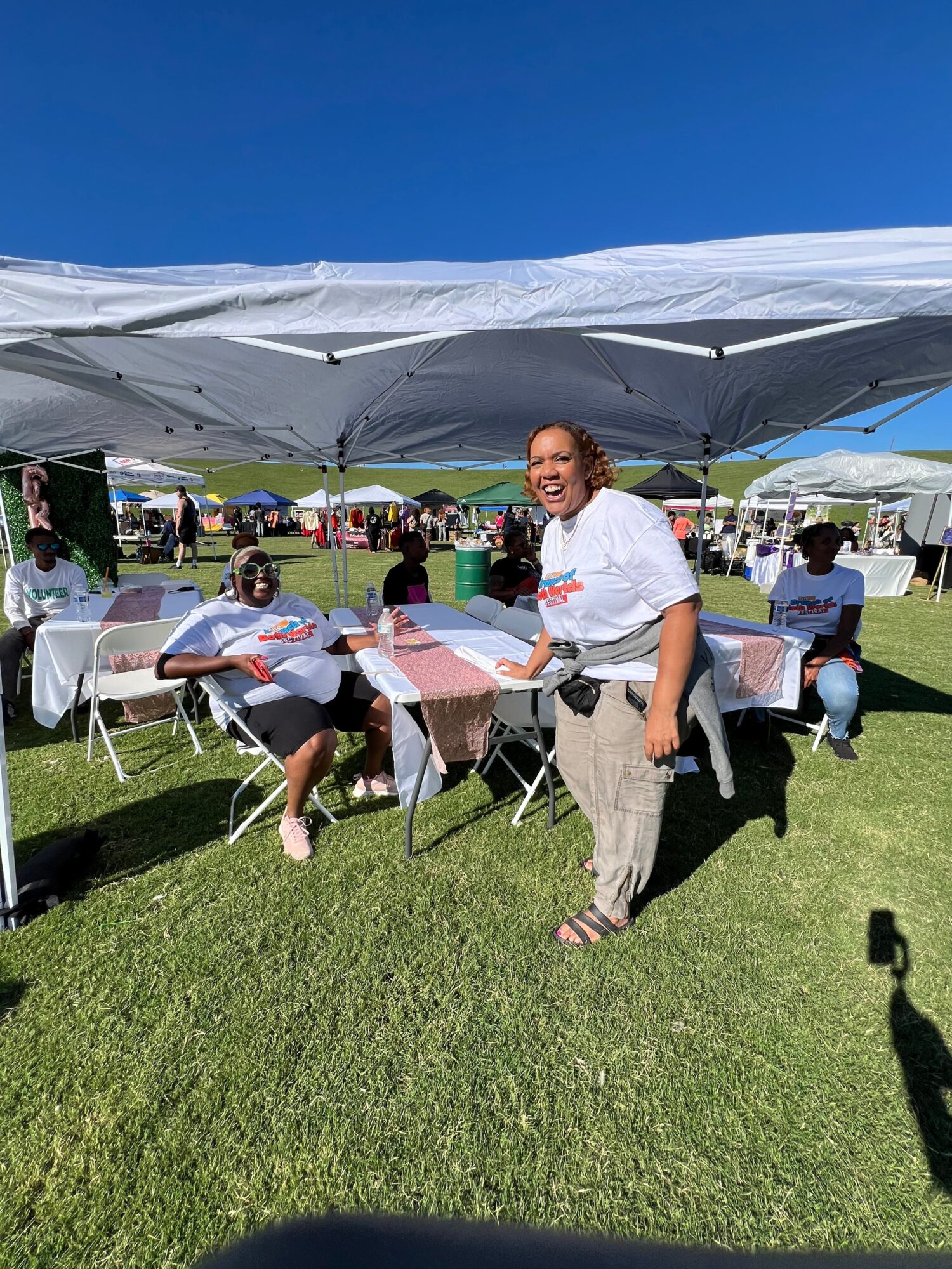 Person speaking at outdoor event with tables and people on grass, under a blue sky.